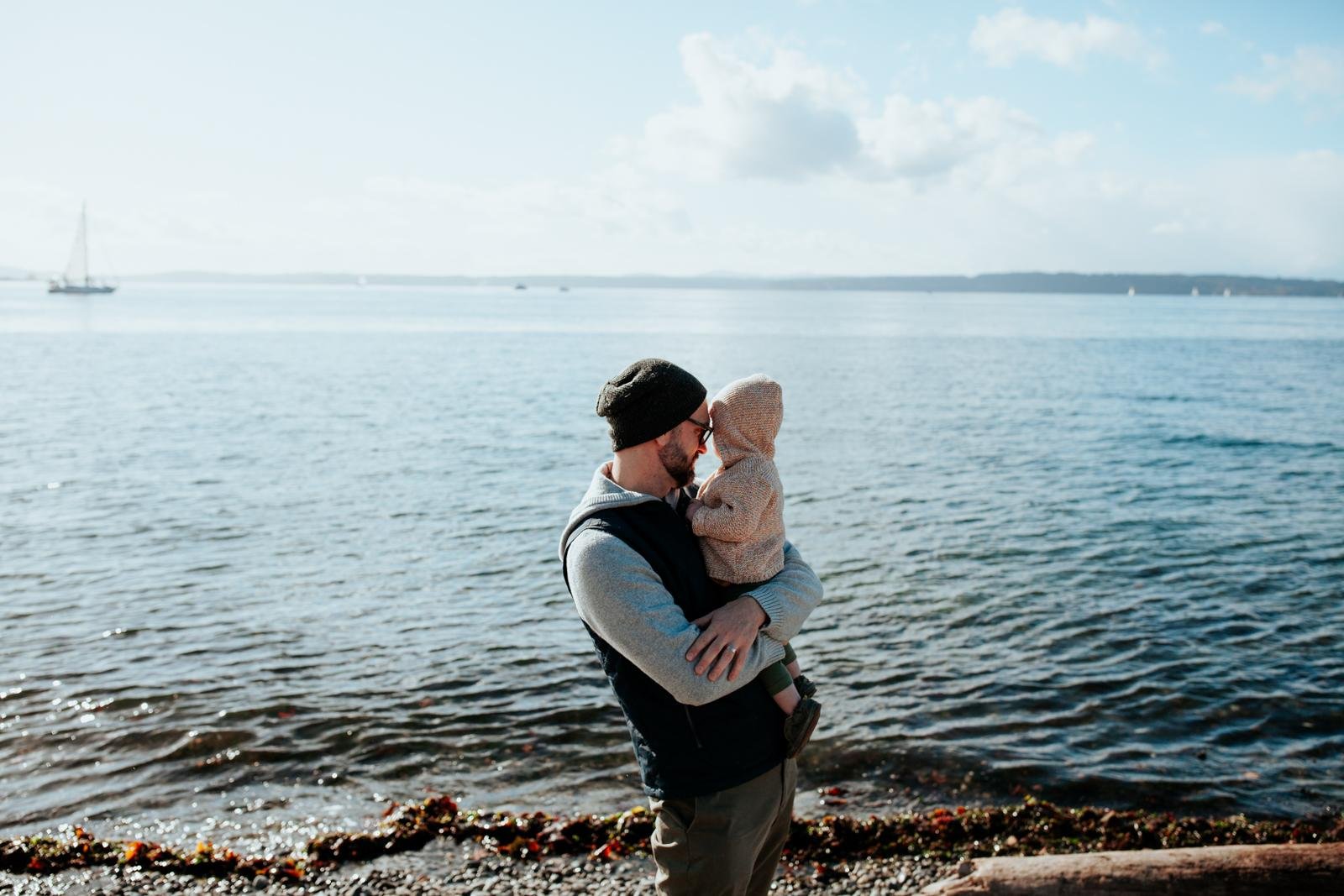  A dad in a black beanie hugs his daughter on a rocky beach for a candid family portrait for Golden Gardens mini sessions in Seattle.&nbsp; 