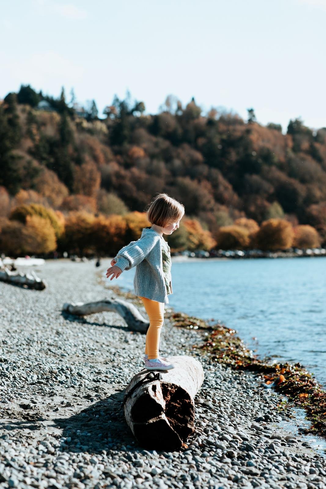  A girl in yellow pants stands on a driftwood log with her arms outstretched on a rocky beach for a candid family portrait for Golden Gardens mini sessions in Seattle.&nbsp; 