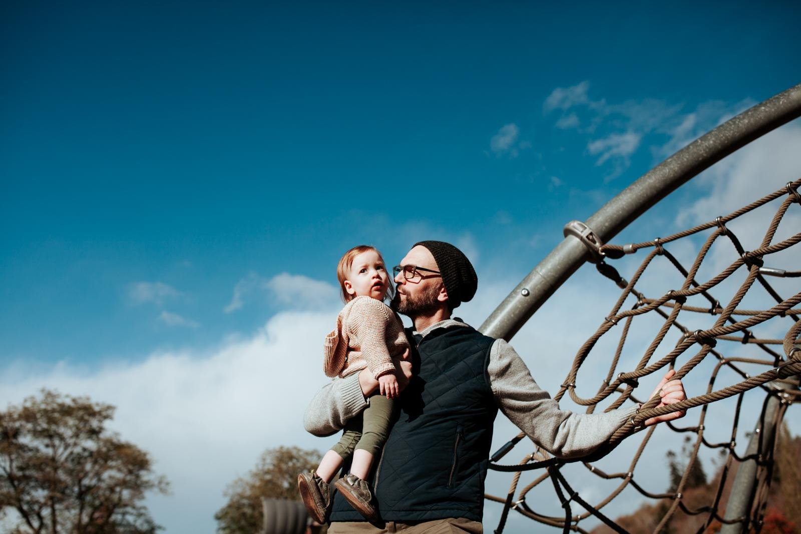  A dad in a black beanie and glasses hugs his baby on a play structure at the park for a candid family portrait for Golden Gardens mini sessions in Seattle.&nbsp; 