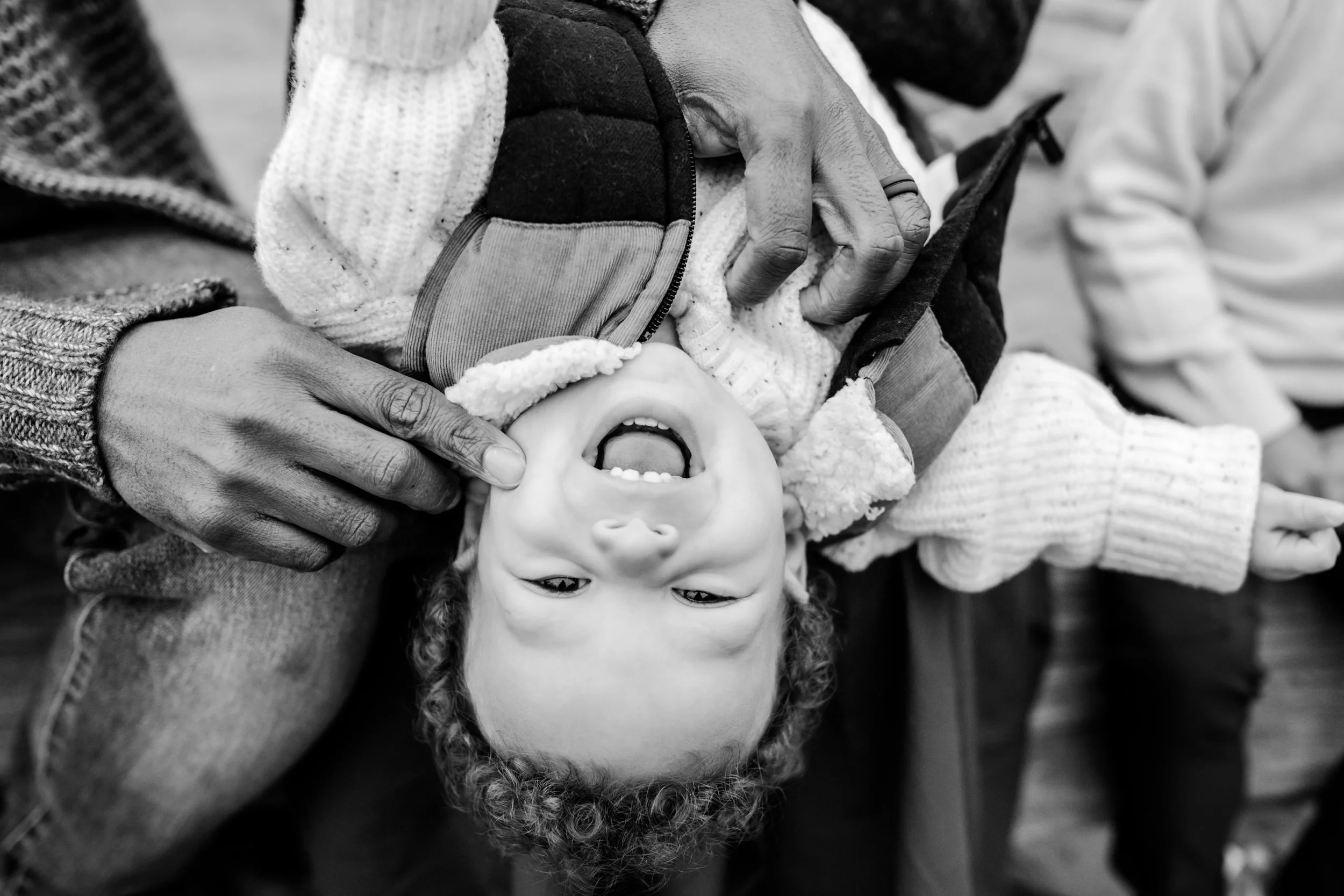 a baby with curly hair smiles while dad plays with him on the beach