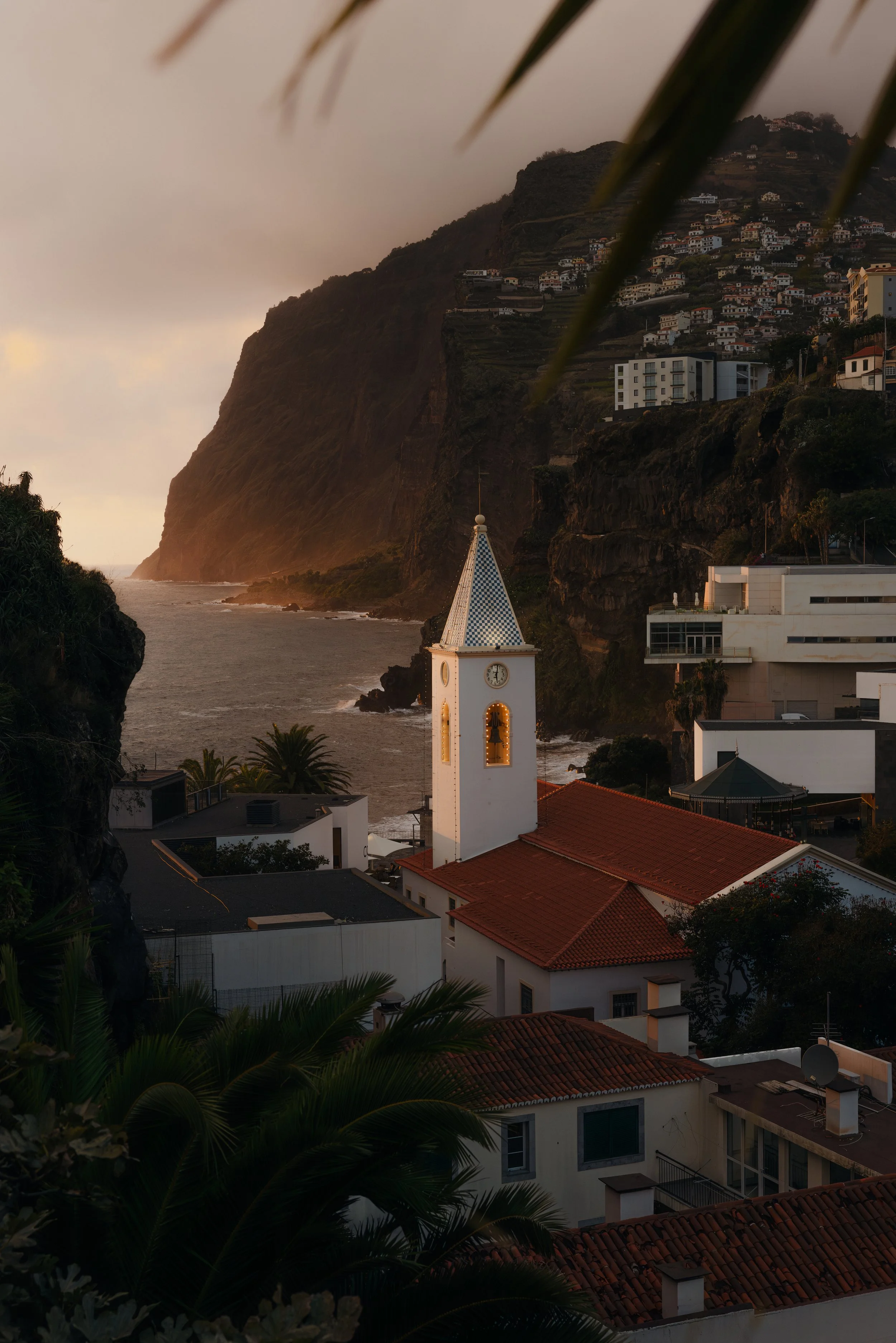 The white bell tower of a church in Câmara de Lobos, Maderia, overlooking a coastal village with dramtic sea cliffs and the ocean at sunset