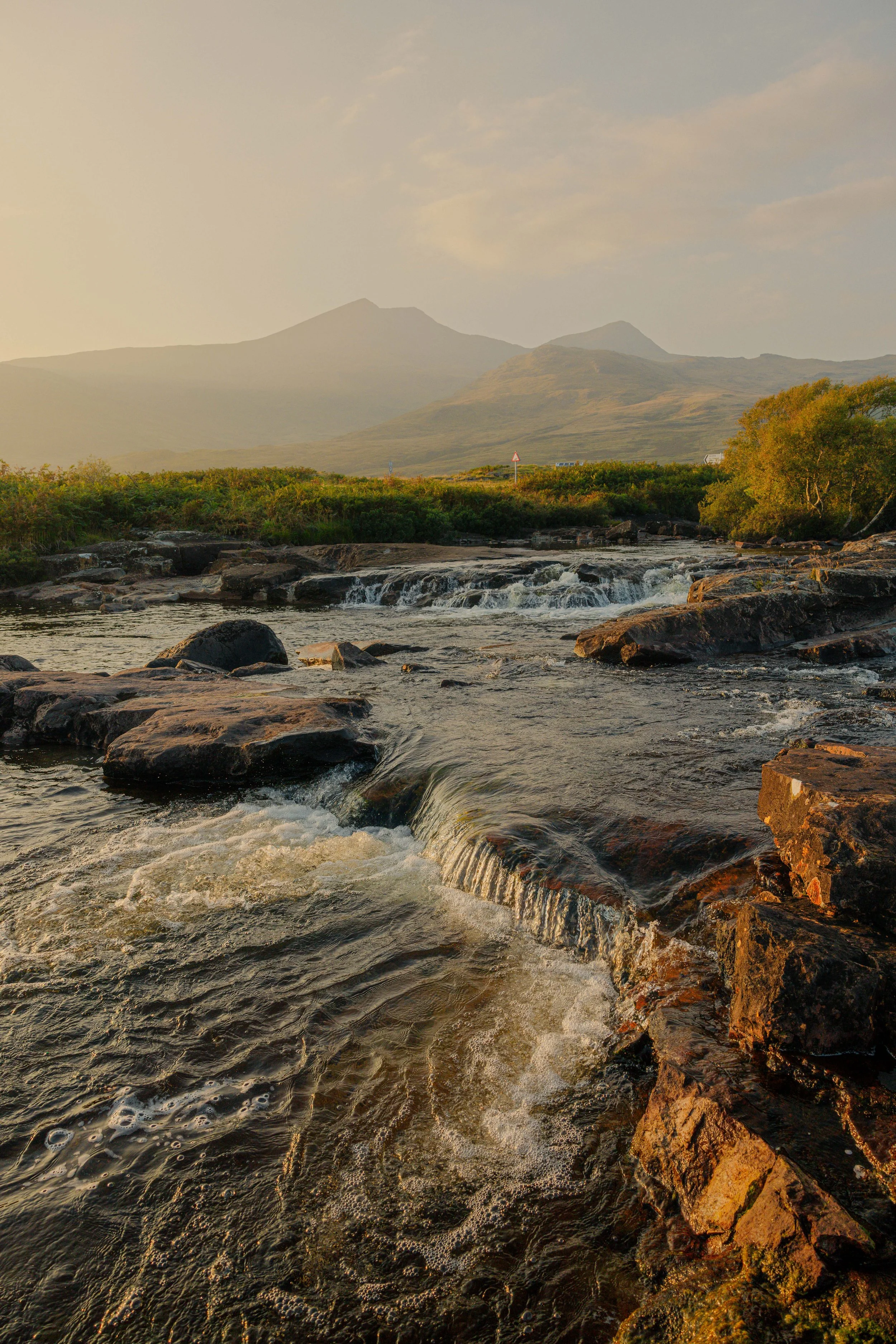 A small waterfall flowing over dark river rocks with a backdrop of Ben More under a hazy summer sky in the Scottish Hebrides