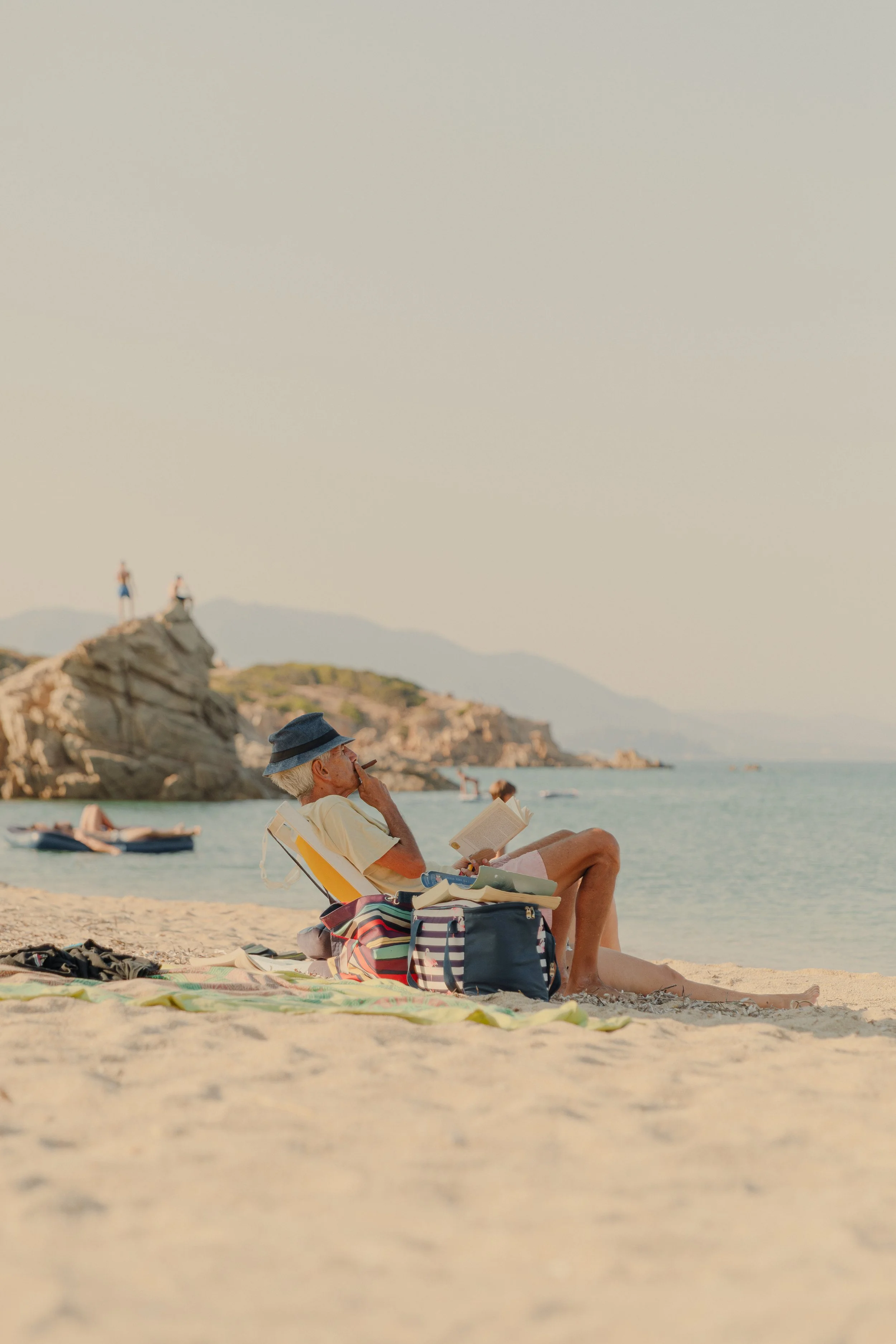 A man in a sun hat is sitting on a beach in Sardinia reading a book and smoking a cigar on a sandy beach. There is a blue sea and rocky coastline in the background