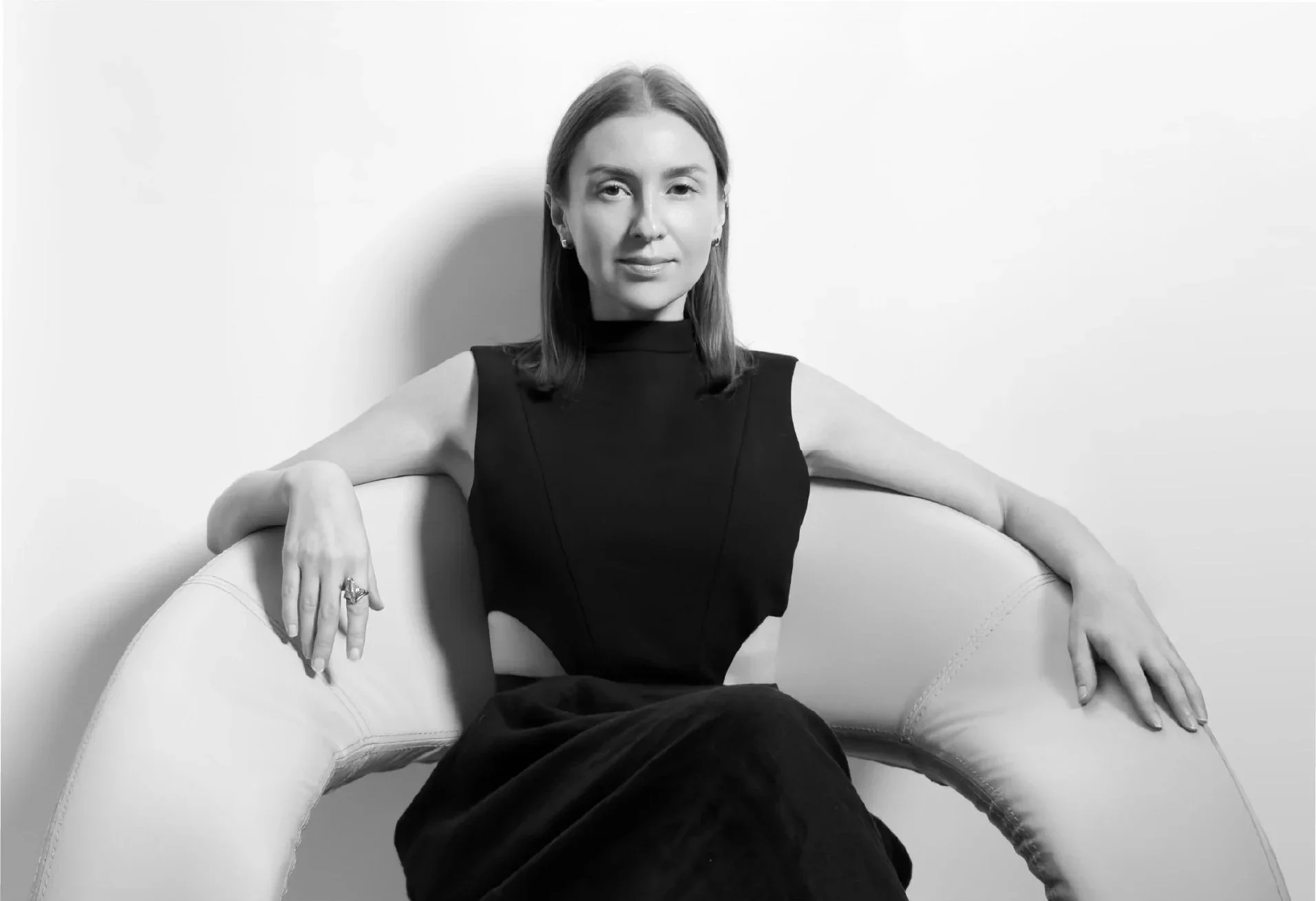 Black and white photo of  Irina Samkova sitting on a curved chair against a plain background, with one arm resting on the chair's back and the other on its side.