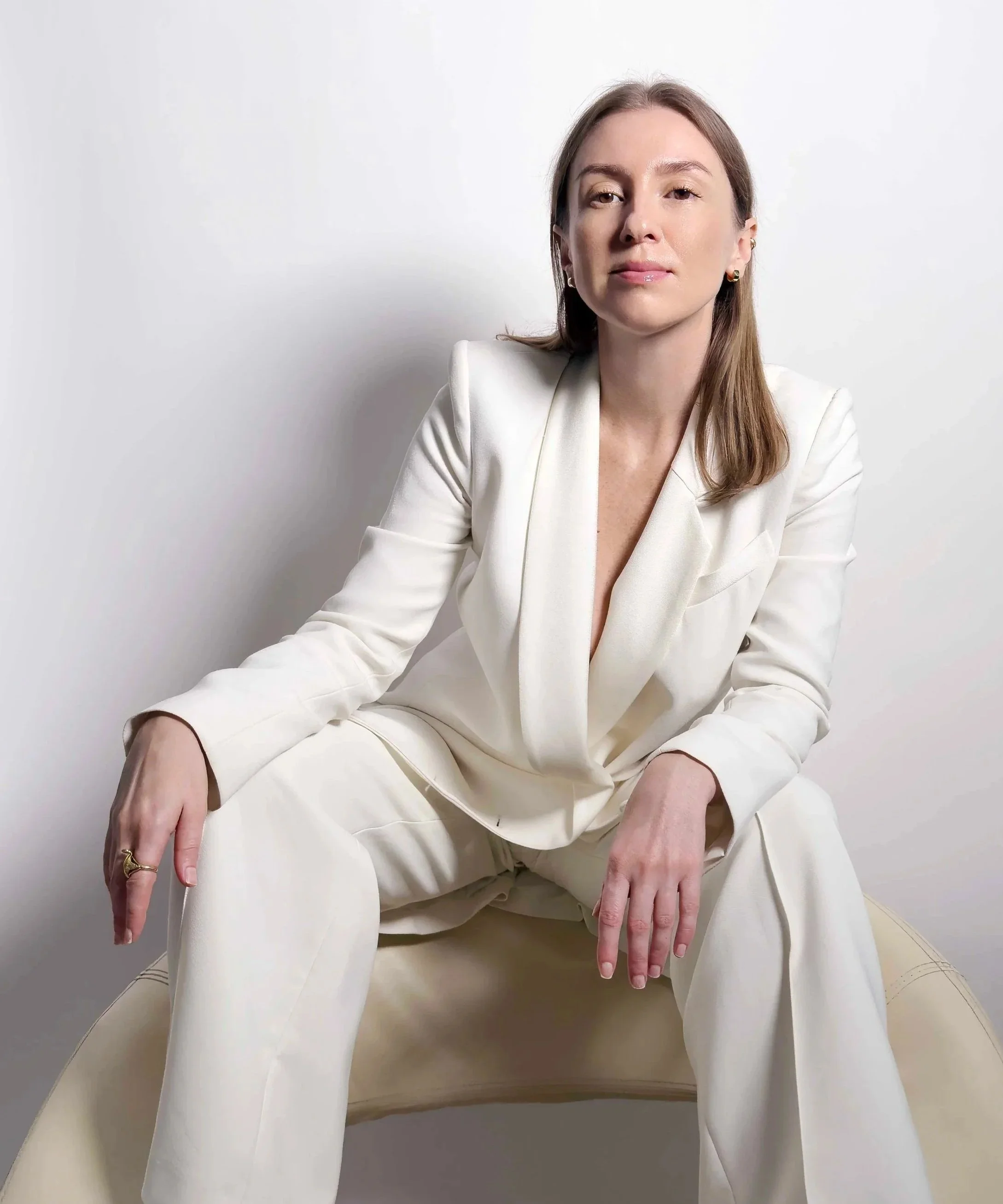 Irina dressed in a white suit, sitting on a white leather chair against a plain white background.