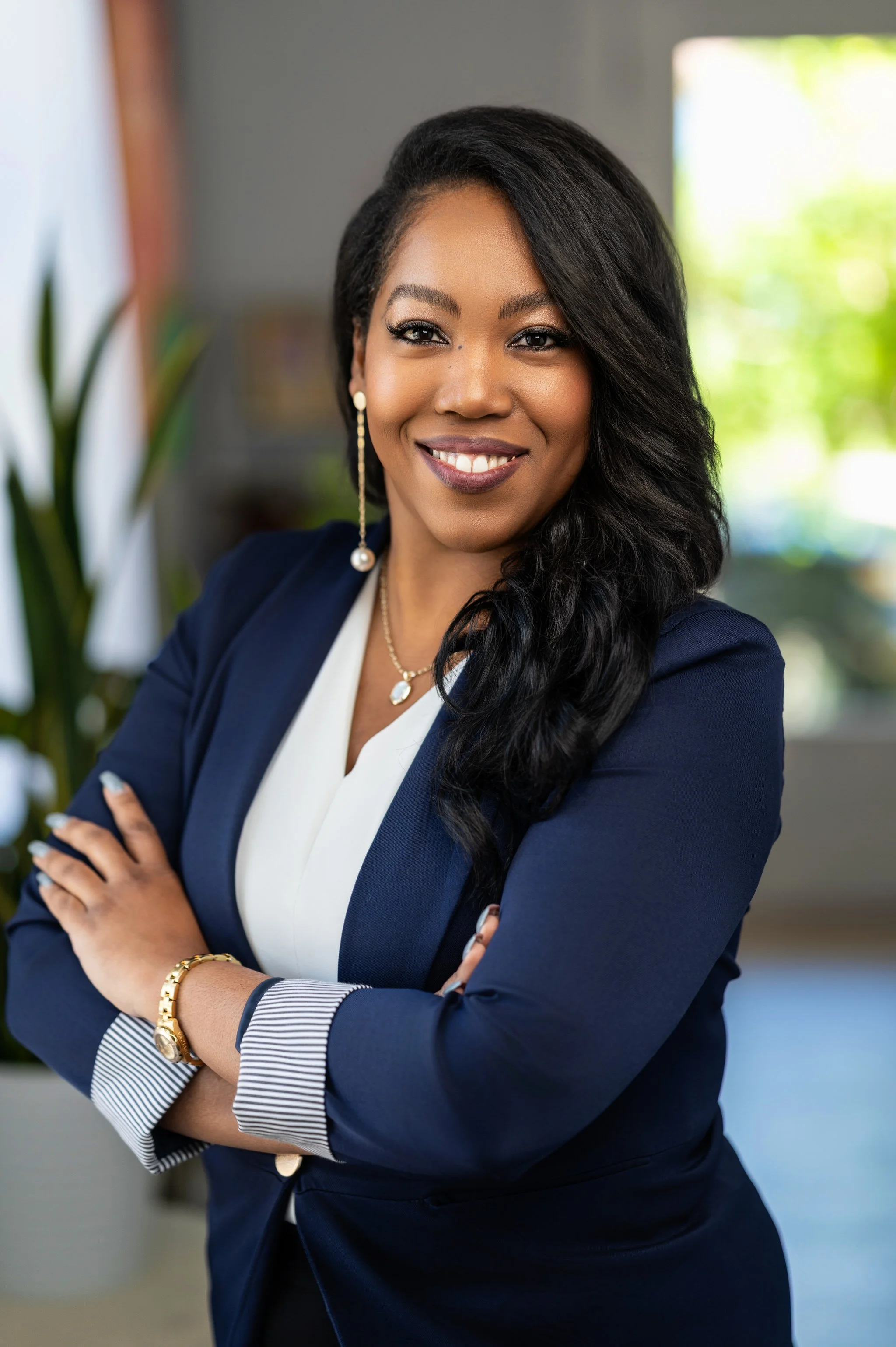 A professional woman with long black hair and dark skin, smiling, wearing a navy blazer and white blouse, standing with crossed arms in an office or home setting with a blurred background and sunlight through window.