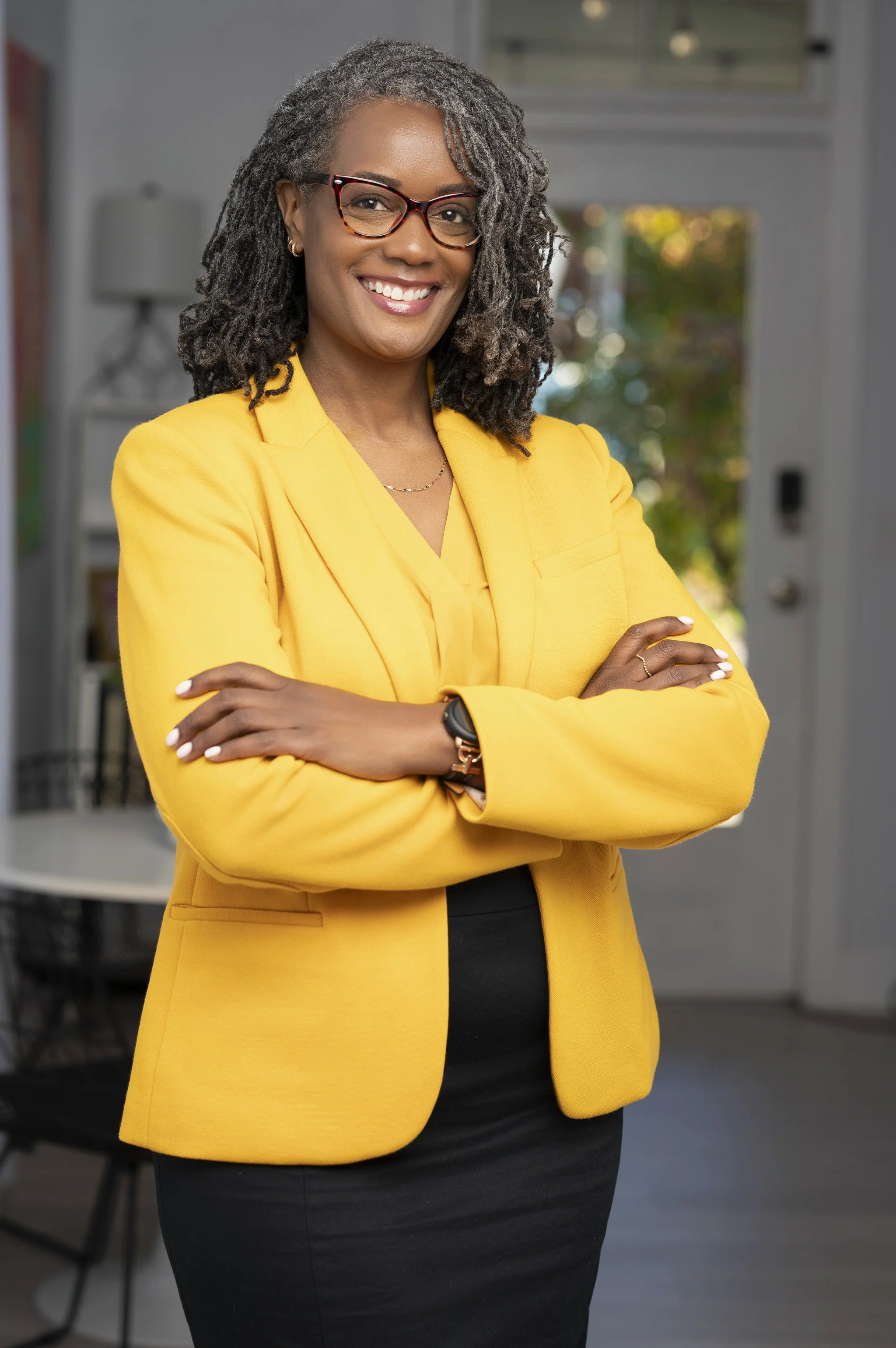 A confident woman stands indoors with her arms crossed, wearing a bright yellow blazer, black pants, and glasses, smiling at the camera.