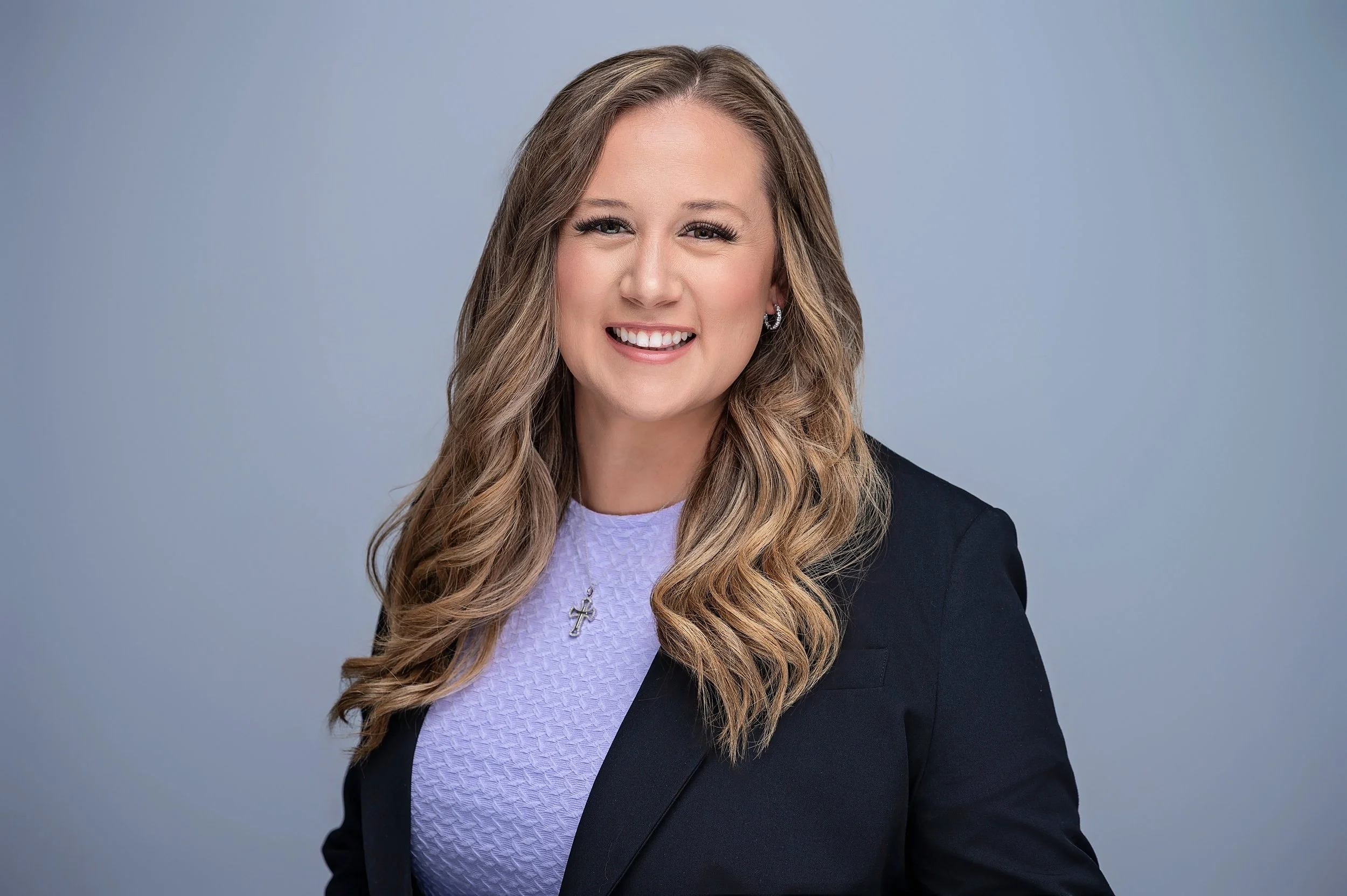 Professional woman with long wavy hair smiling, wearing a black blazer, light purple textured top, and a cross necklace, against a gray background.