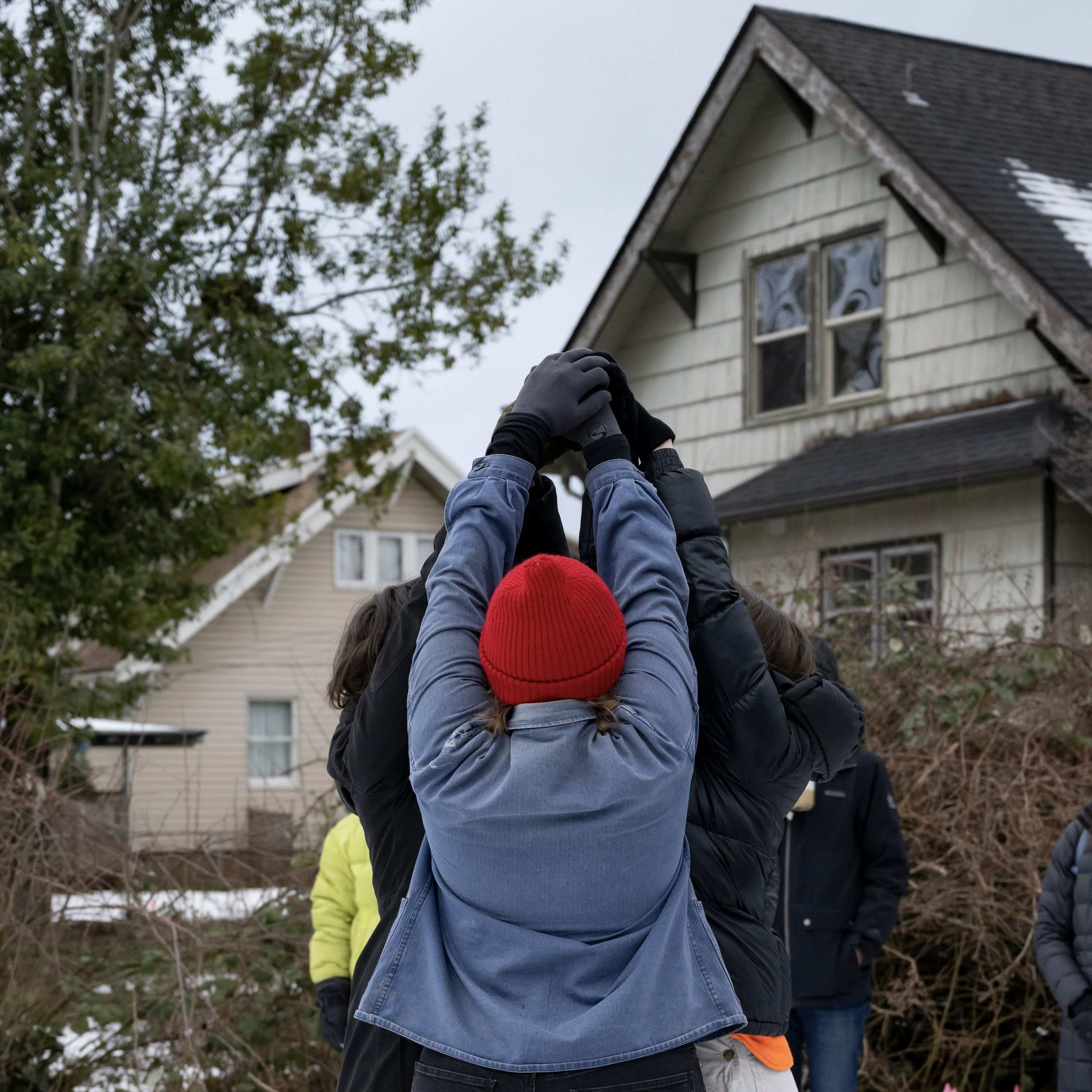  three people’s arms reaching up together to the sky to create a pyramid shape on a snowy grey day. They are in the backyard, and the eaves of a house are visible behind them.  