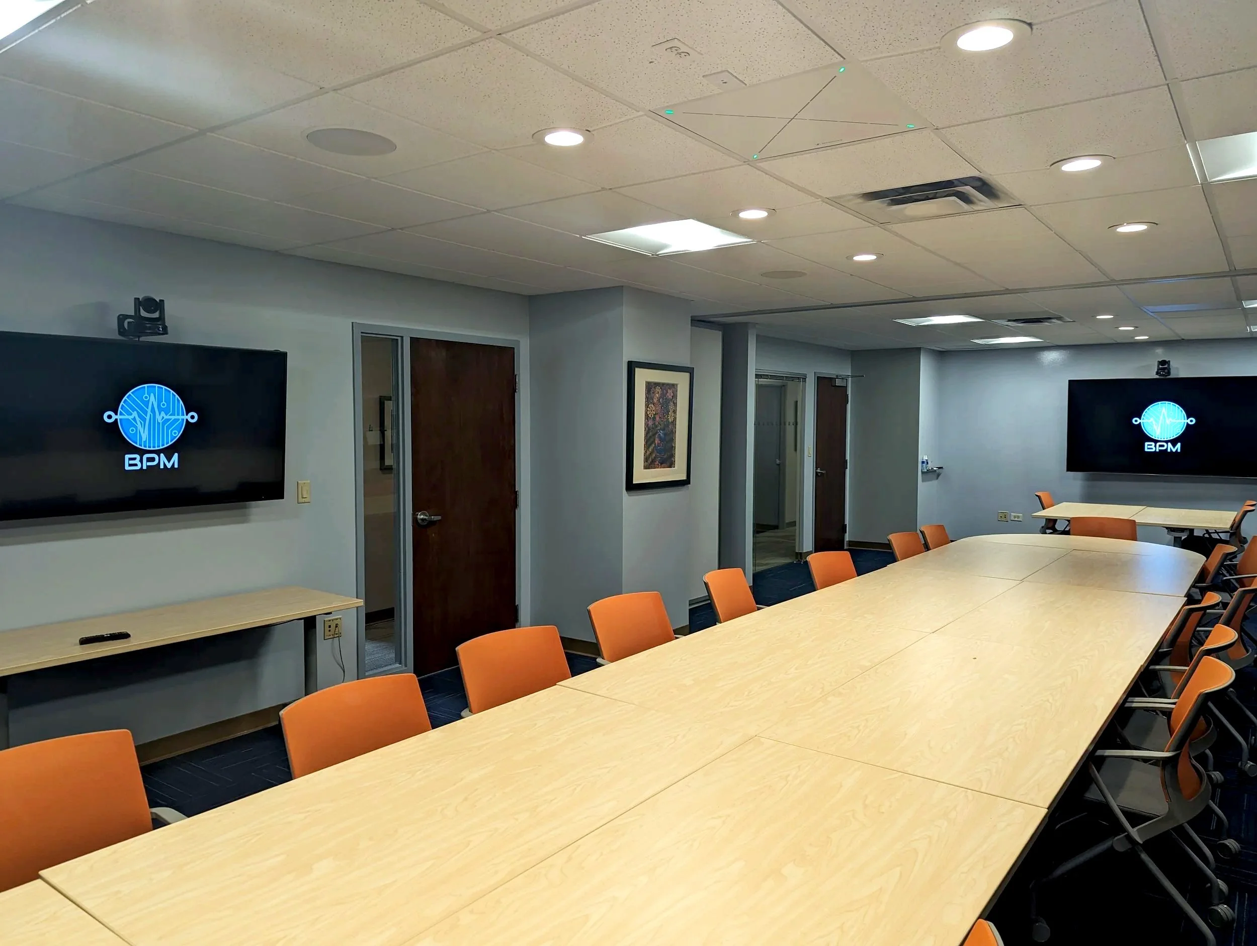 A conference room with a long light wooden table surrounded by orange chairs, mounted flat-screen TVs displaying a blue digital logo with lines and the text 'BPM' on a gray wall, ceiling lights, framed picture, and doors in the background.