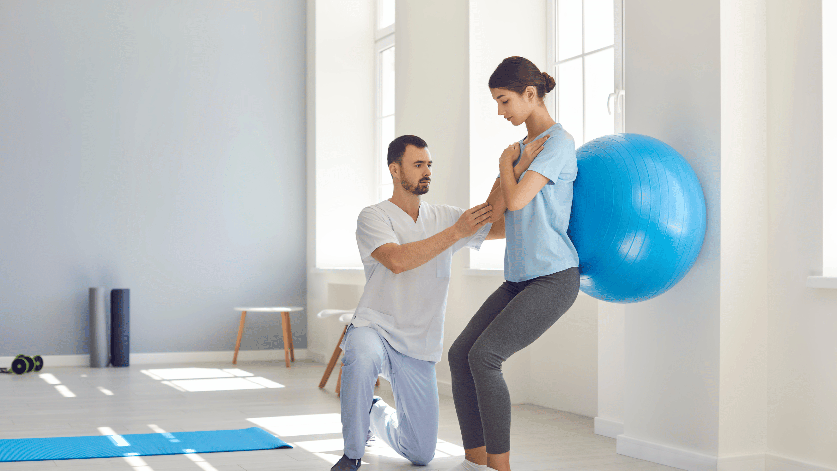 Physical therapist supporting female patient leaning against yoga ball and wall.