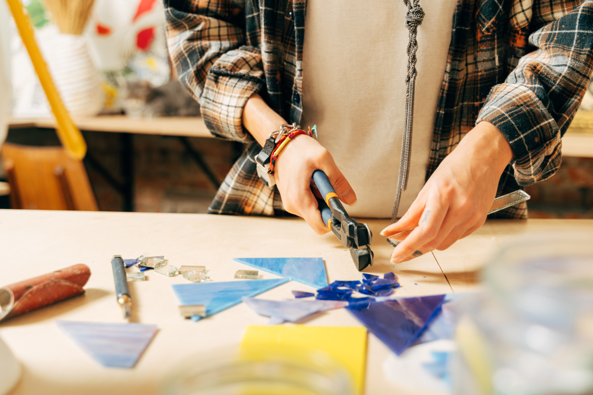 Person wearing a flannel shirt, holding the proper tools to work with blue-colored glass pieces