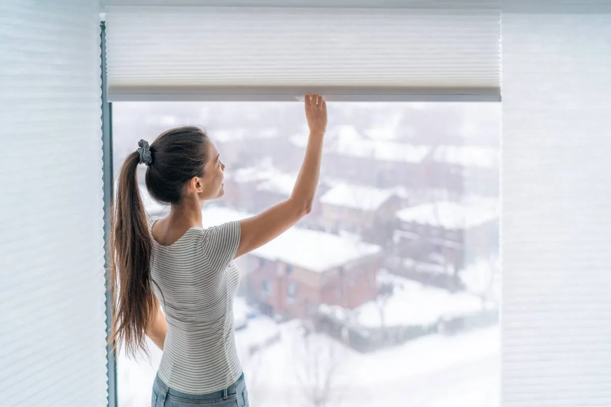 A woman opening her cellular shades to look at the snowy view outside her window