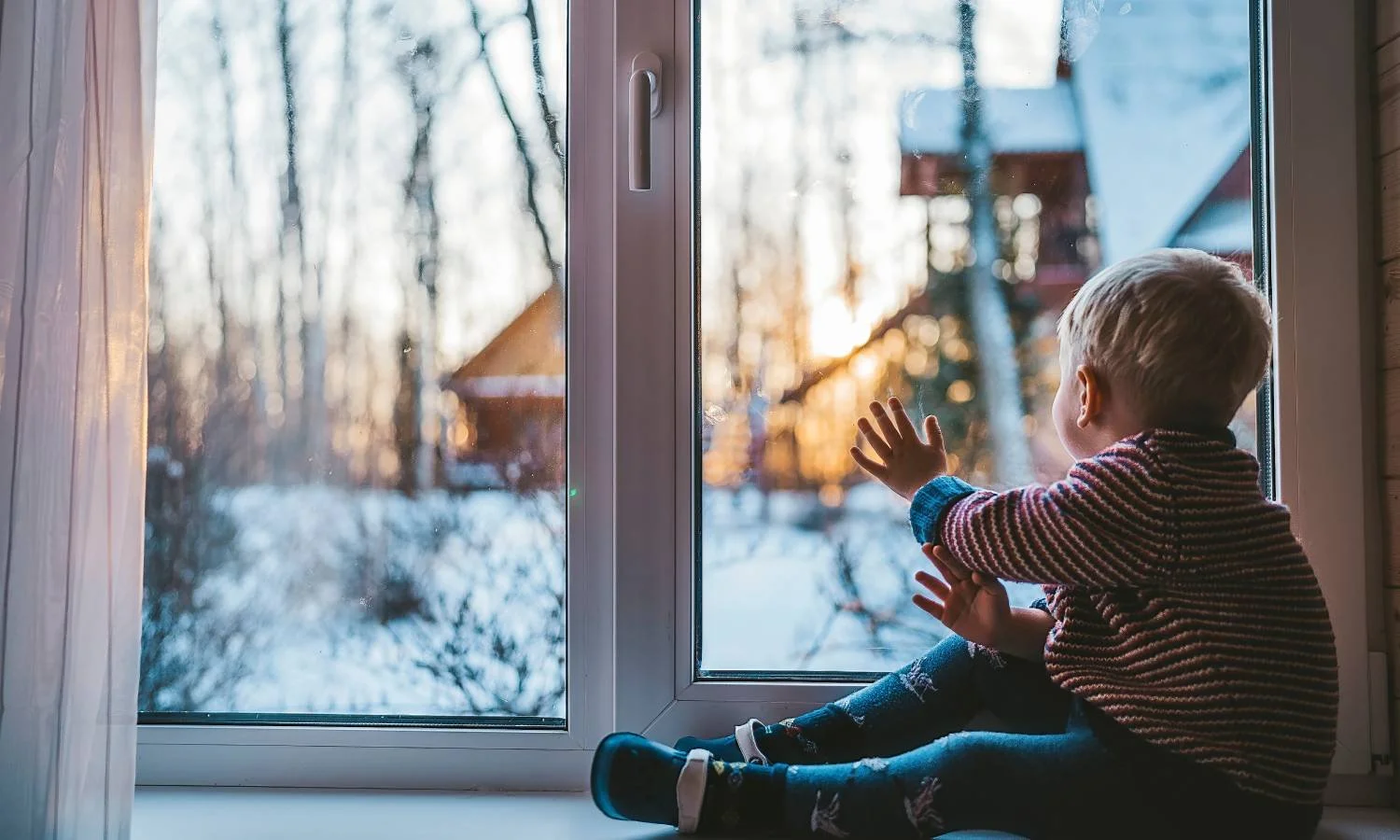 A little boy sitting with his hand on a custom storm window, looking out at snow