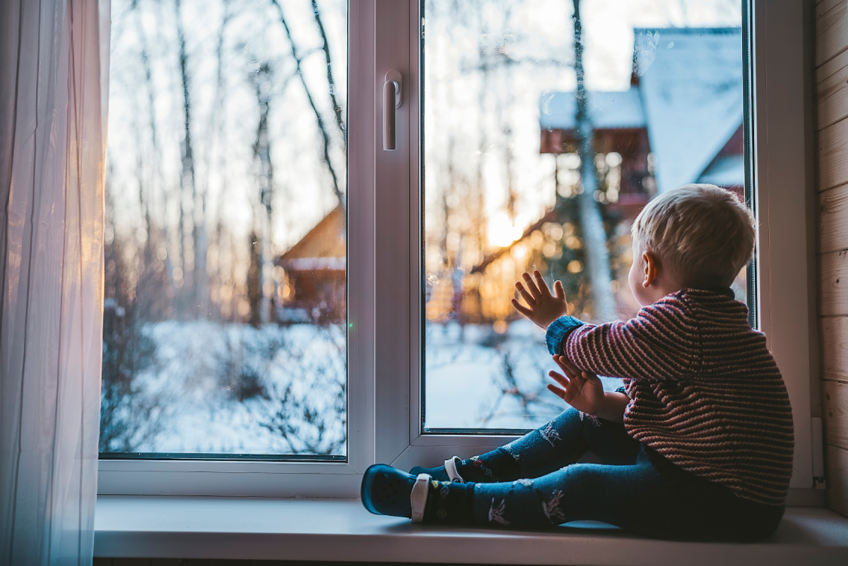 A little boy sitting with his hand on a custom storm window, looking out at snow