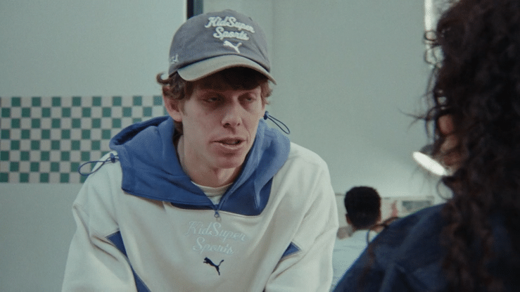 A young man wearing a gray baseball cap with 'KidSuper Sports' logo, a blue hoodie, and a white jacket with a Puma logo, talking to a woman with curly dark hair in what appears to be a fast-food restaurant or diner.