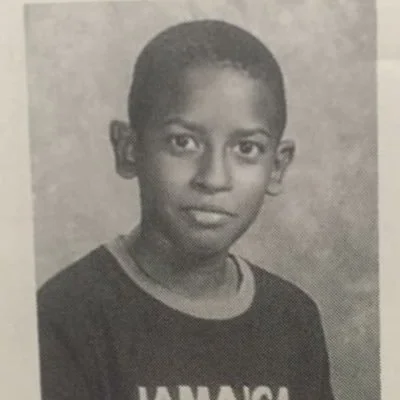 A black and white portrait of a young boy with short hair, wearing a dark shirt with partially visible text.