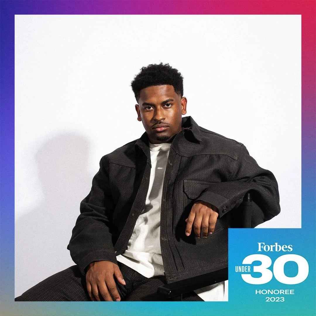 A young man with short curly black hair, wearing a black jacket over a white shirt, sitting against a plain white background. There is a blue overlay with the words "Forbes Under 30 Honoree 2023" at the bottom right corner.