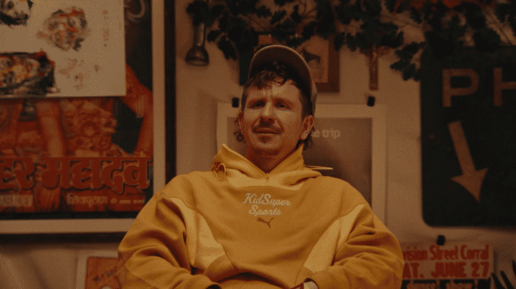 A man with dark hair and a slight mustache, wearing a yellow hoodie and a gray cap, sitting indoors with a somewhat confused or serious expression.