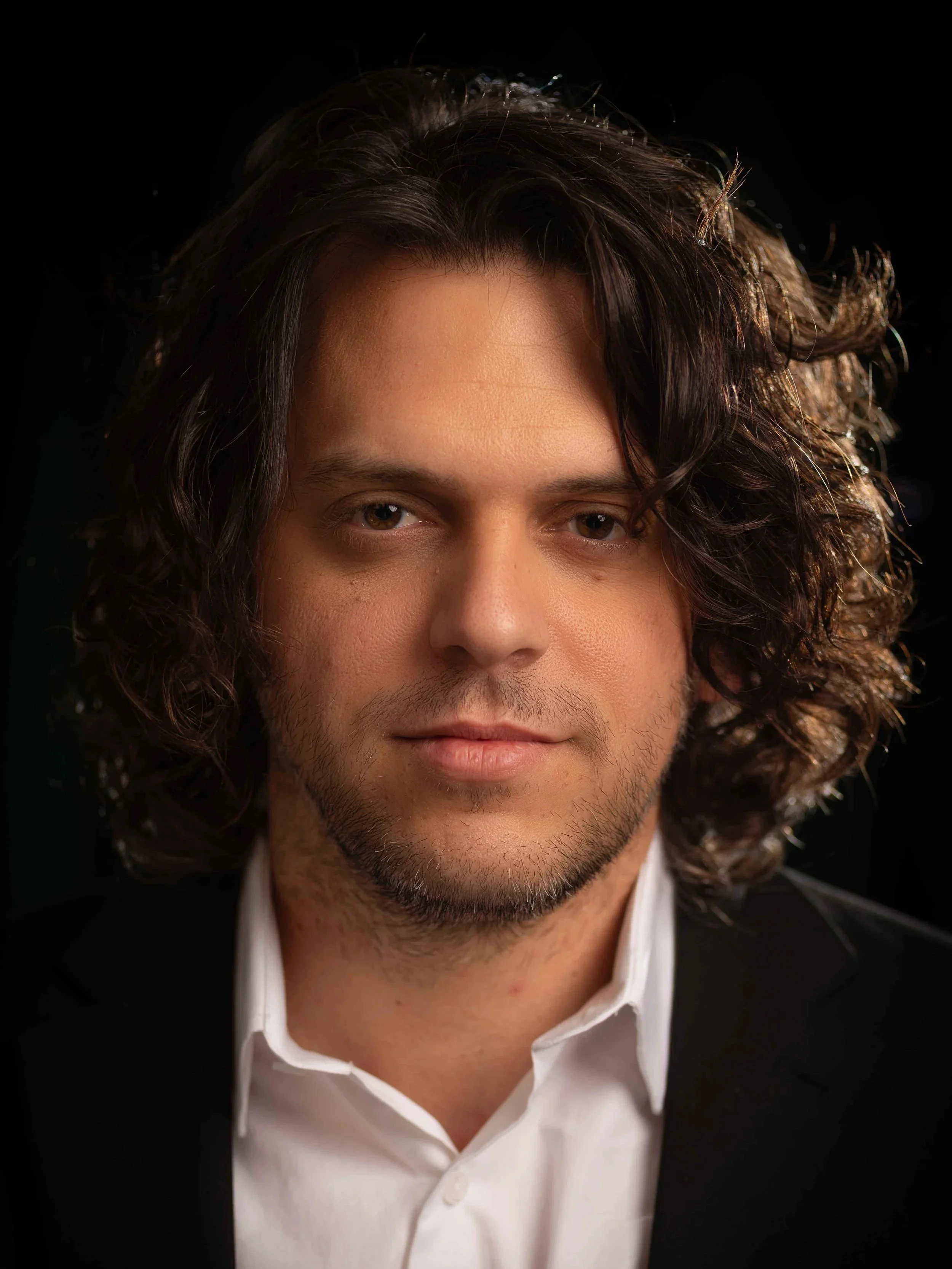 A smiling man in a black t-shirt sitting at a sound mixing console in a recording studio.