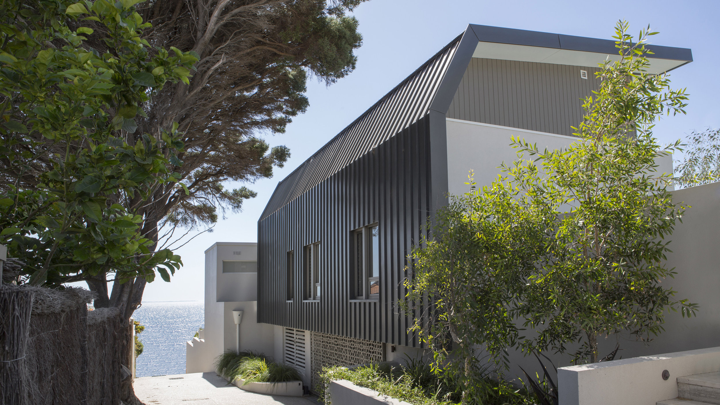 Beach St. The project was centred on the concept of converting an upside-down home (living upstairs) into a home with a conventional connection to external spaces. The solution was unconventional and involved the raising of the external spaces to the rear by 3m to access the existing living spaces, creating a greater level of transparency through the existing home and creating a new (fourth level) to access the panoramic views from the roof. This solution presented numbers of complex approval and construction hurdles that are the unseen foundation of this complex project. These included overlooking, building on boundary, overshadowing, excavation, retaining and height issues. The home including a number of complexities and challenges in the integration and harmonisation of the existing 3 storey home with a new 3 storey addition, resulting in a 4 storey residence. A careful balance was followed through the maintenance of the existing built form with the new architectural form. The decision was made to complete the home in an integrated and sympathetic palette. This included introducing design elements into the existing home to stitch the two together.