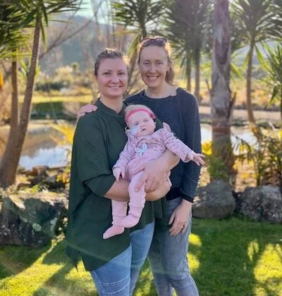 Tania and Gwen smiling, holding Riley, outside in front of a green setting of palm trees and rockery.