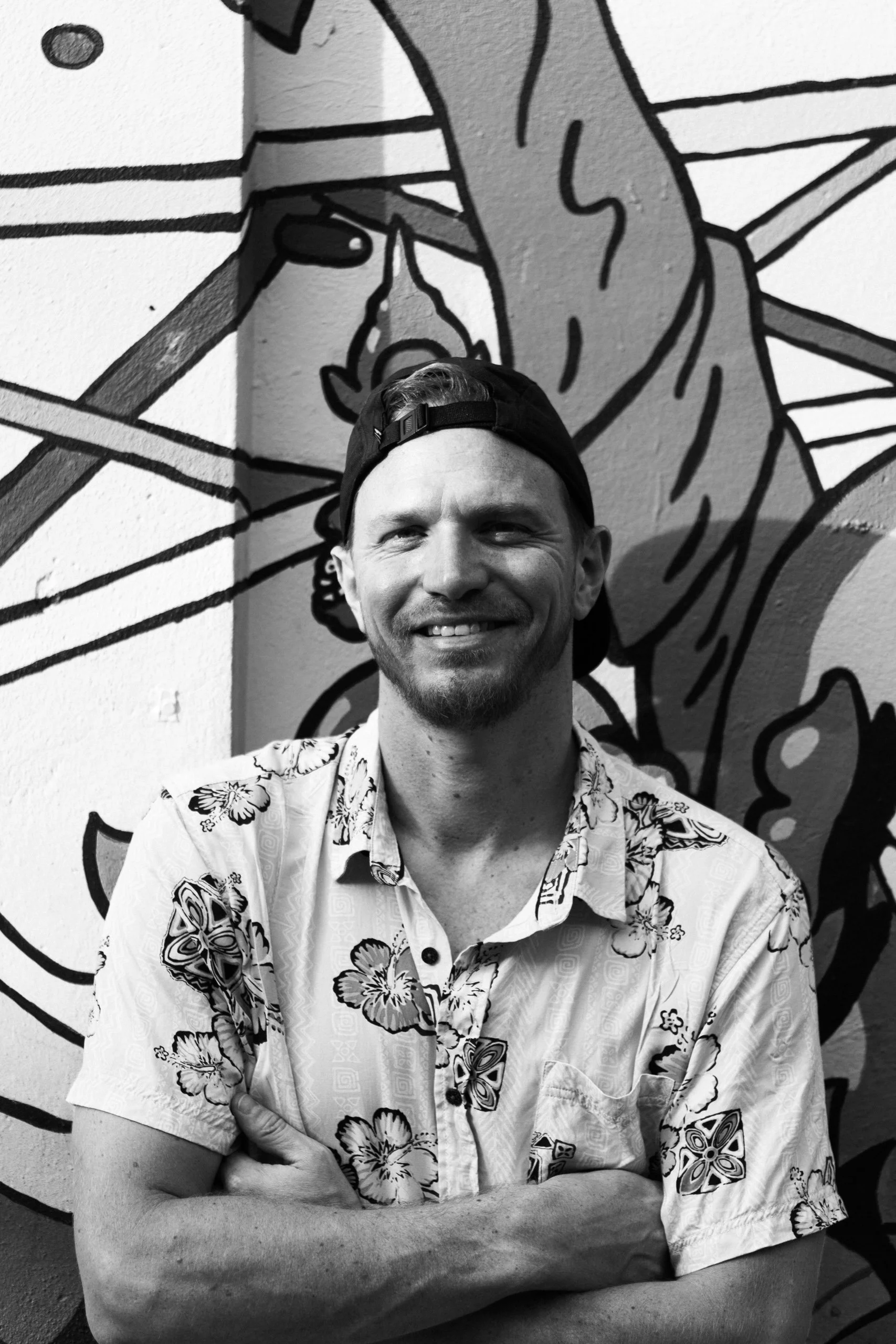 Mann mit Baseballcap und Hawaii-Hemd steht vor einer Wand mit Straßenkunst, lächelt