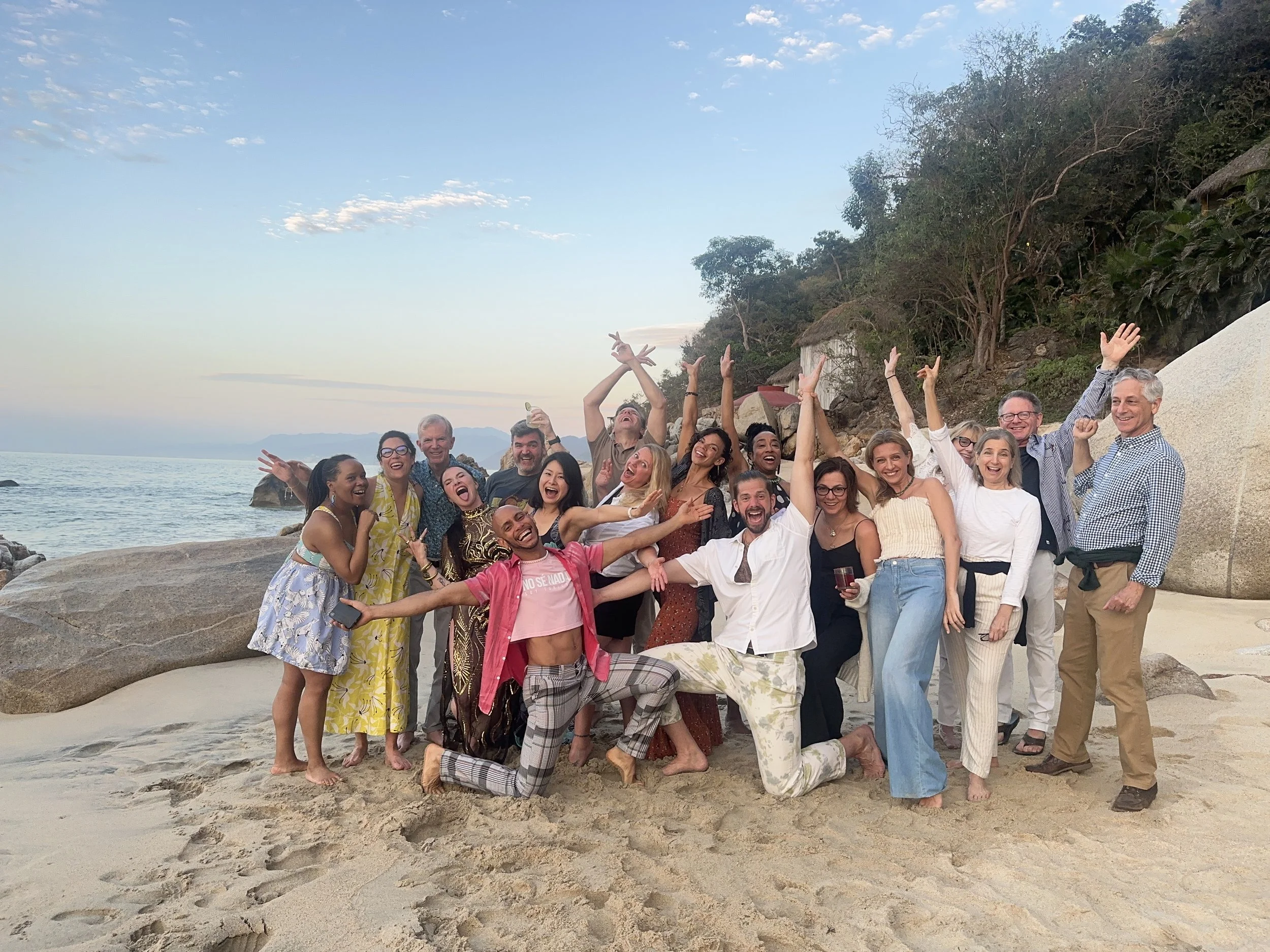A large group of people standing and sitting on a beach with rocks and trees, celebrating and posing for a photo during sunset.