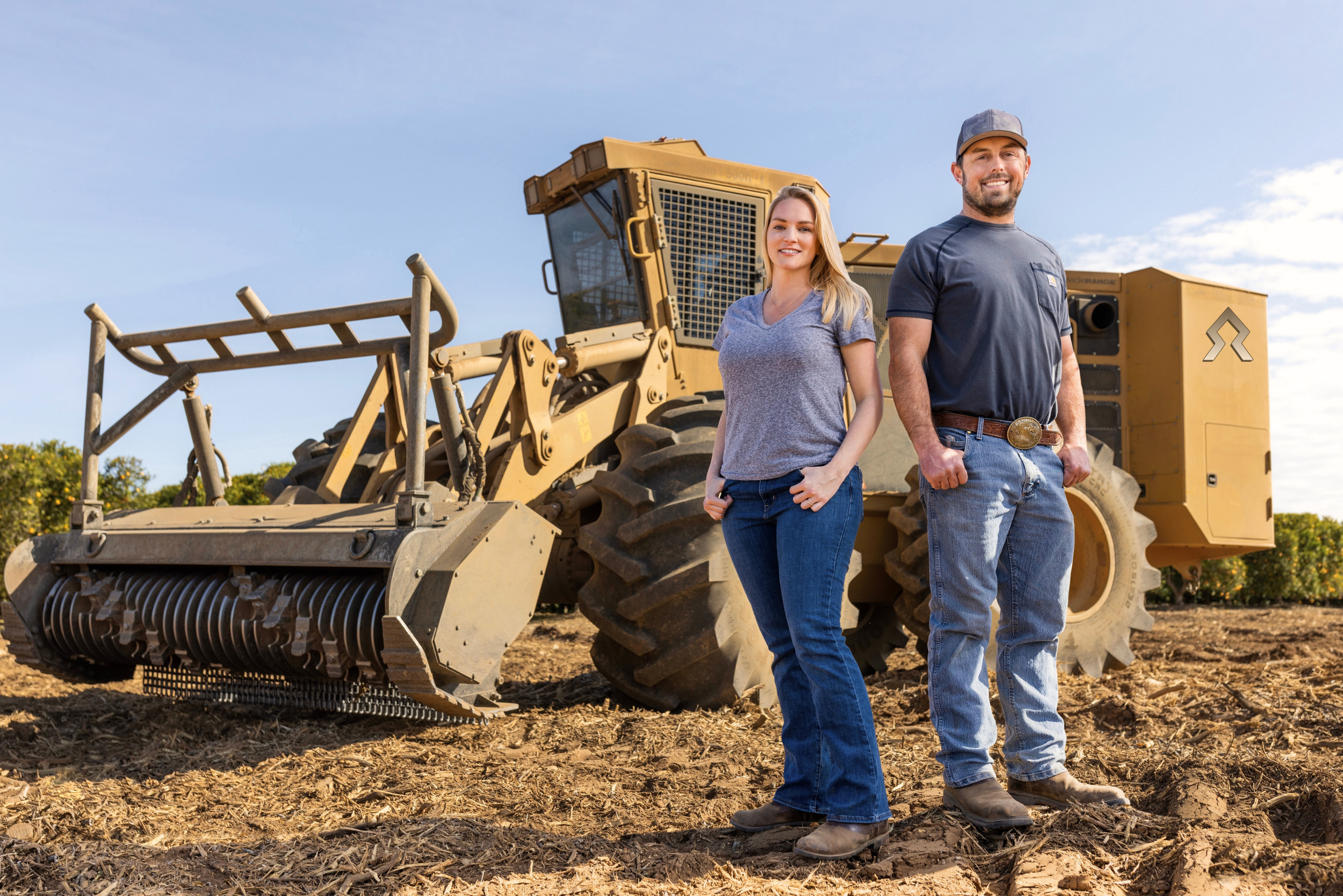 A man and woman standing in front of a large construction vehicle on a dirt field, smiling at the camera.