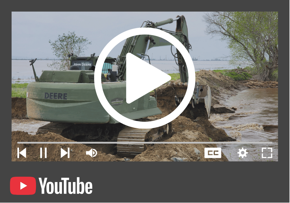A John Deere excavator positioned on a riverbank during daytime, with muddy water and trees in the background.