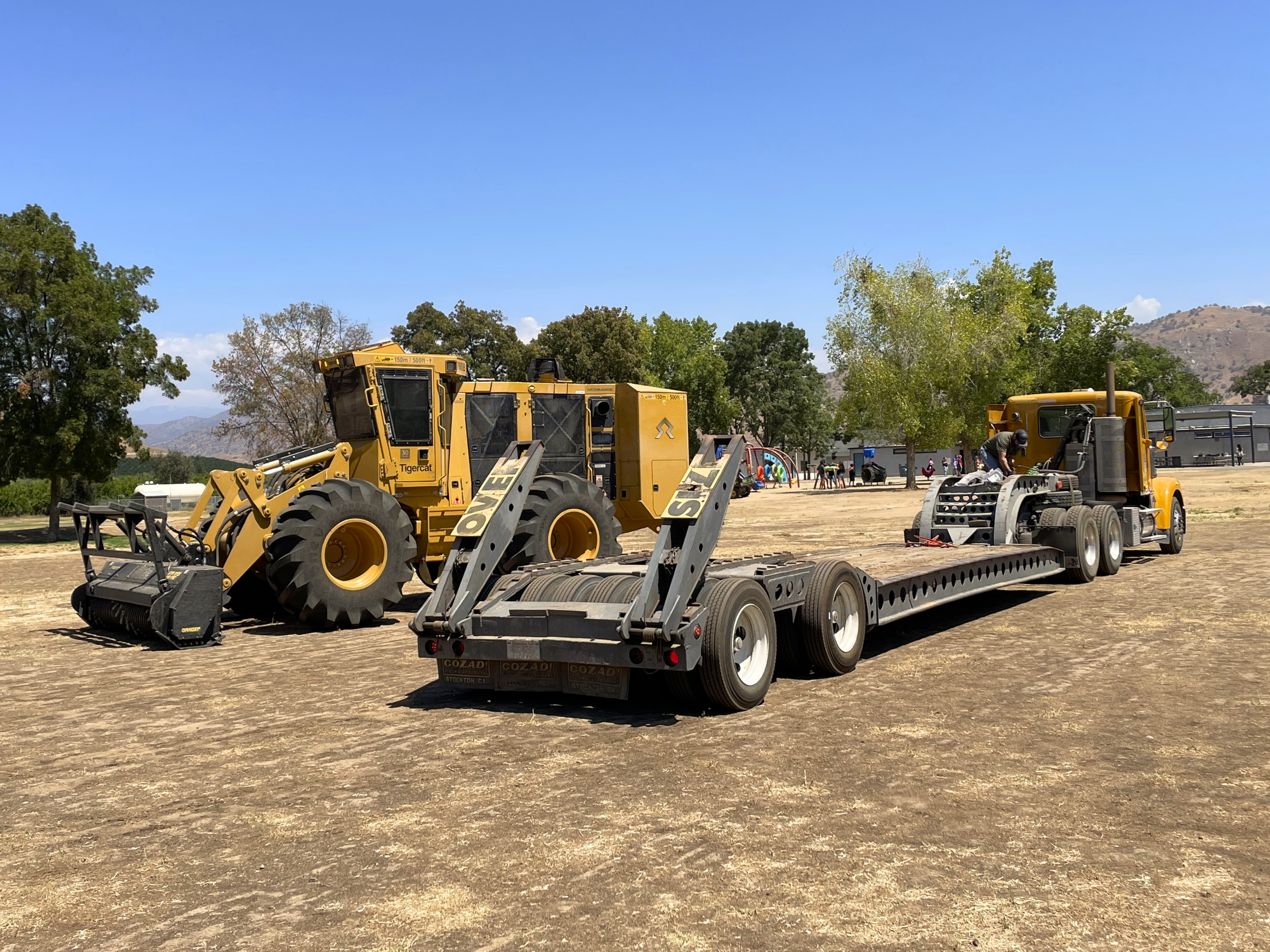 Construction equipment and a flatbed truck in an outdoor area with trees and hills in the background.
