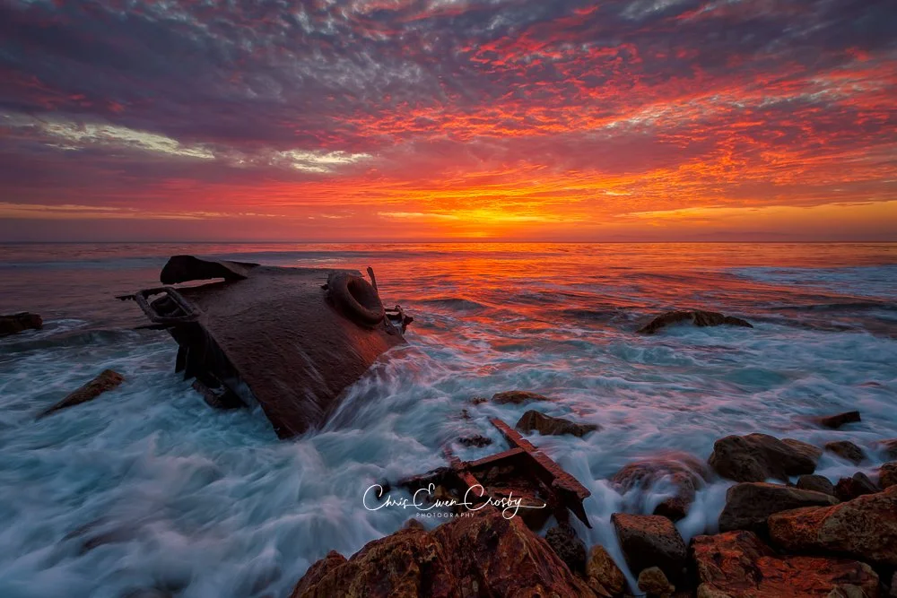 A wide-angle landscape photograph of a rusted shipwreck on a rocky California beach during a vibrant, colorful sunset with swirling clouds.