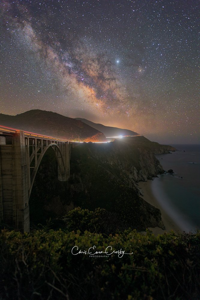Vertical night photograph of Bixby Bridge in Big Sur with car light trails on Highway 1 and a starry night sky above the Pacific coast.