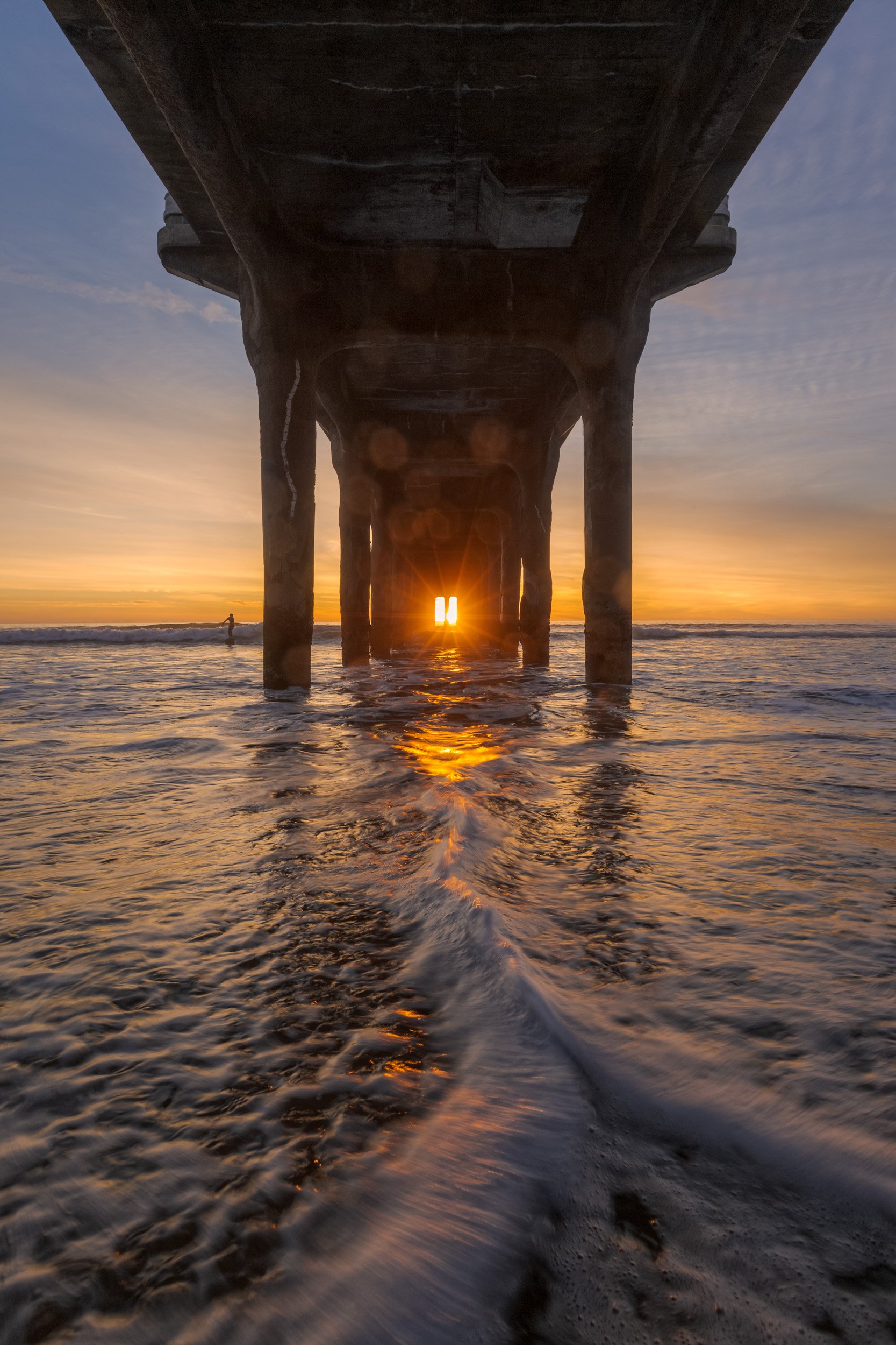 Vertical shot looking under the Manhattan Beach Pier pilings as the setting sun aligns directly in the center with rushing tide water in the foreground by Chris Ewen Crosby.