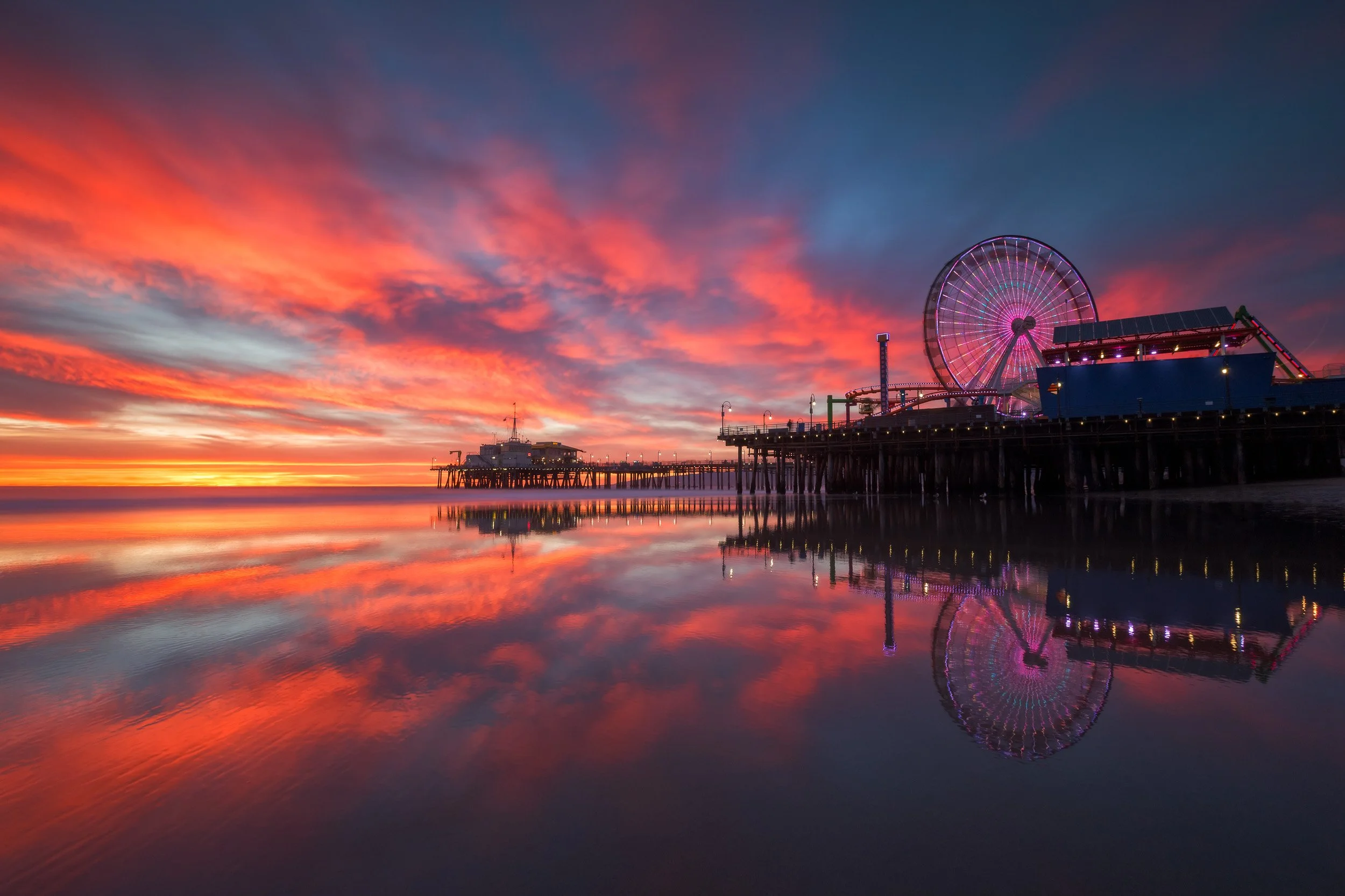 Vibrant fiery orange sunset over the Santa Monica Pier and Pacific Park Ferris wheel reflected on wet shoreline sand by Chris Ewen Crosby.