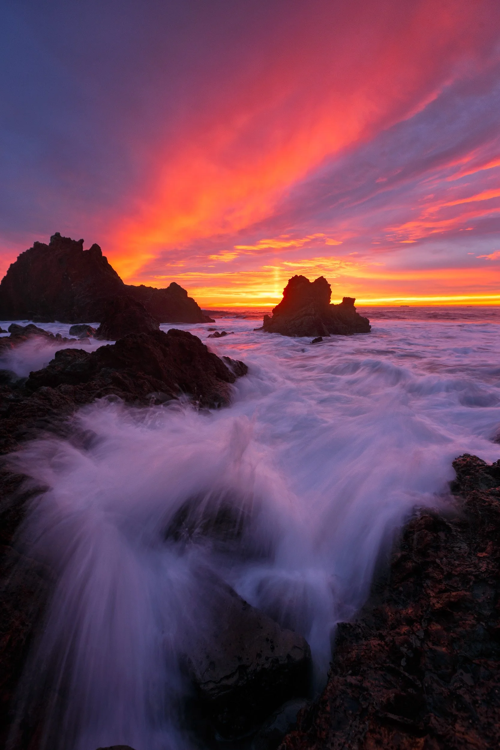 Vertical long exposure photograph of a dramatic crimson and magenta sunset sky over rocky sea stacks and rushing whitewater in Palos Verdes by Chris Ewen Crosby.