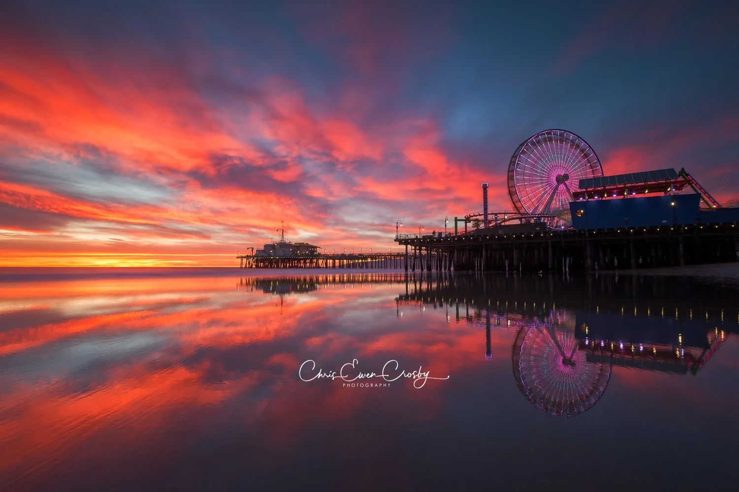 A horizontal landscape photograph of Santa Monica beach at sunset, with fiery orange and purple clouds reflecting perfectly on the wet, glassy sand.