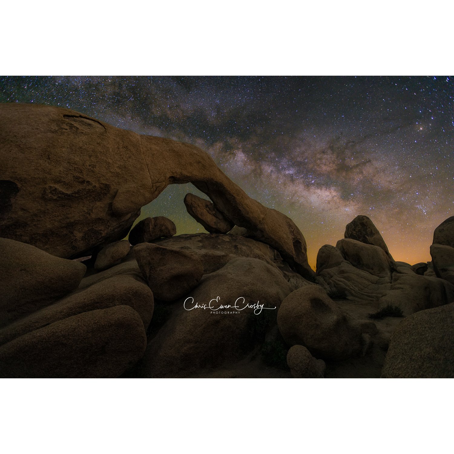 A horizontal landscape night photograph of a natural stone arch in Joshua Tree National Park with the bright Milky Way galaxy core visible in the starry sky above.