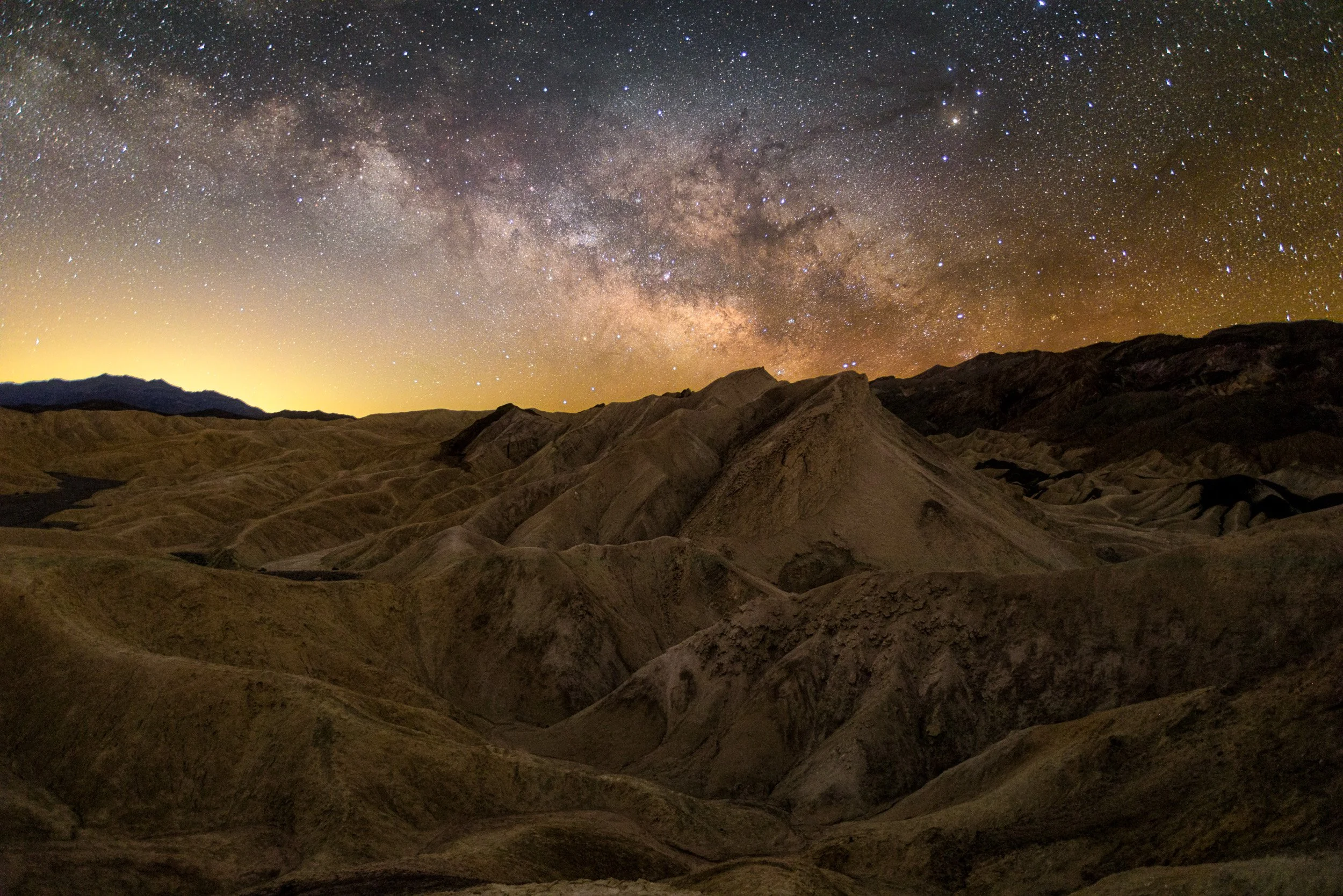 Horizontal night astrophotography photograph of the golden, eroded badland ridges at Zabriskie Point illuminated beneath the Milky Way galaxy core in Death Valley National Park by Chris Ewen Crosby.