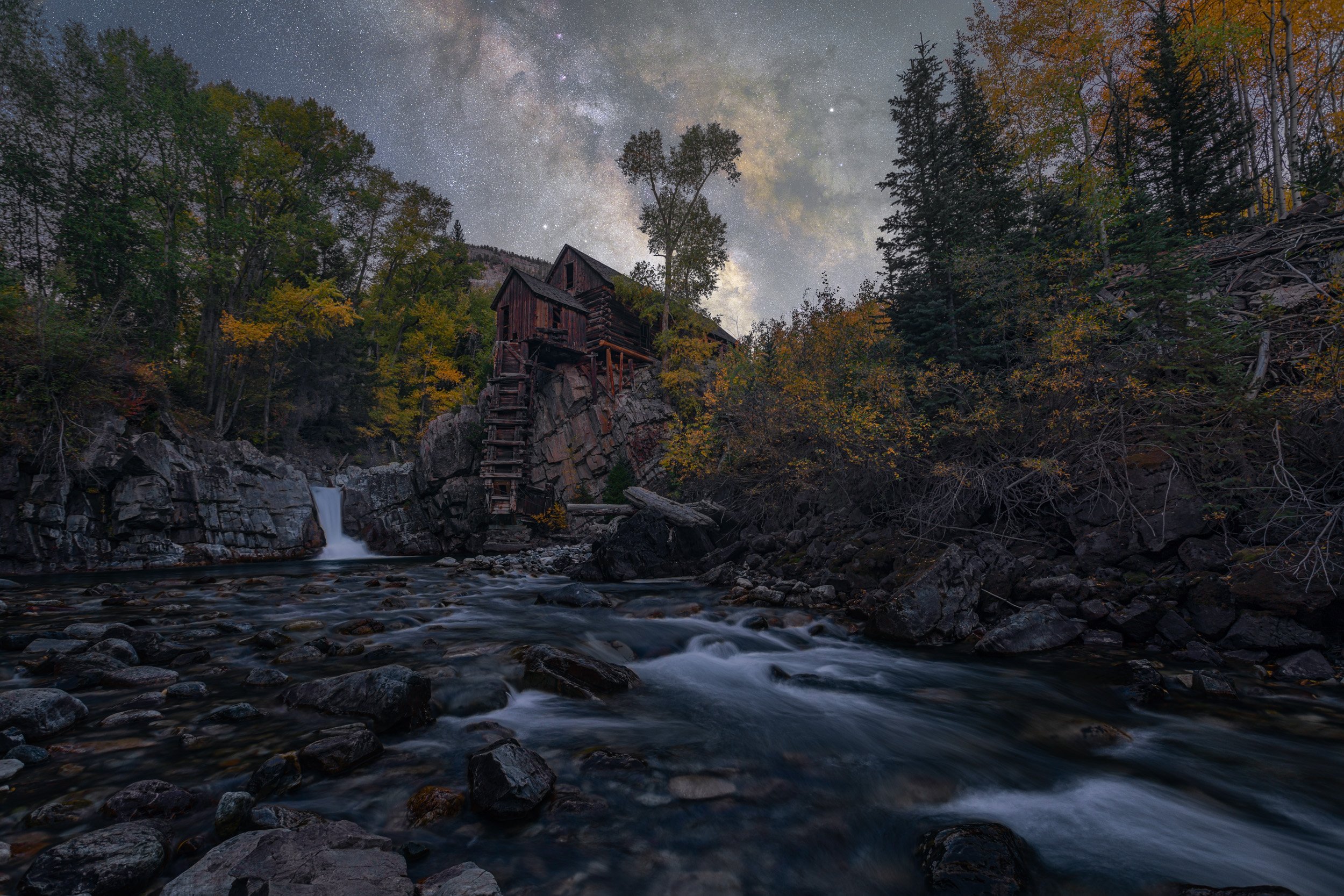 Horizontal night astrophotography photograph of the historic Crystal Mill surrounded by autumn foliage and the rushing Crystal River under the glowing Milky Way in Marble, Colorado by Chris Ewen Crosby.
