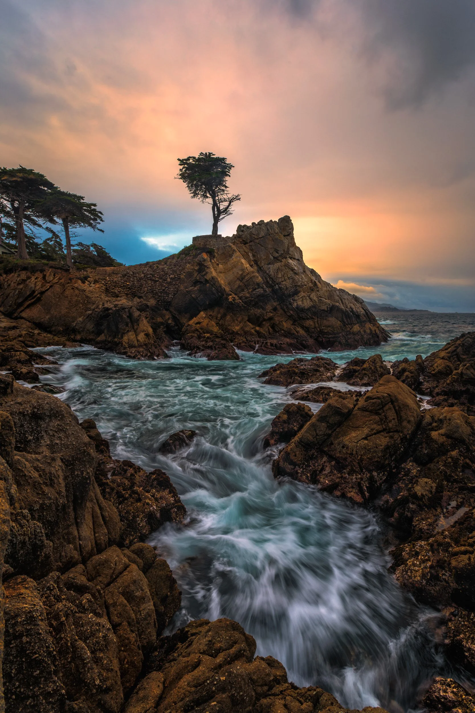 Vertical photograph of the iconic Lone Cypress tree perched atop a rugged granite cliff above turquoise ocean waters under a dramatic sunset sky in Pebble Beach by Chris Ewen Crosby.