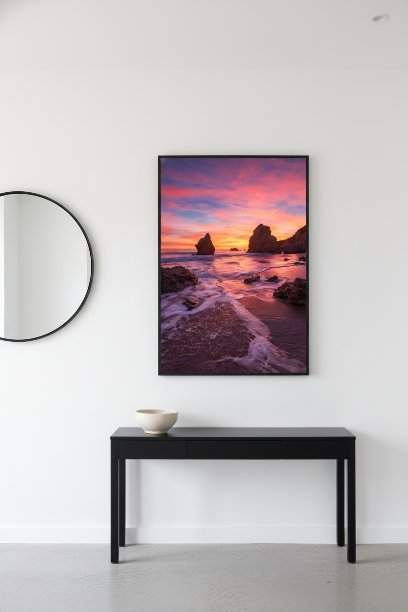 Framed El Matador Beach sunset photo print mounted above a black entryway console table next to a round mirror.