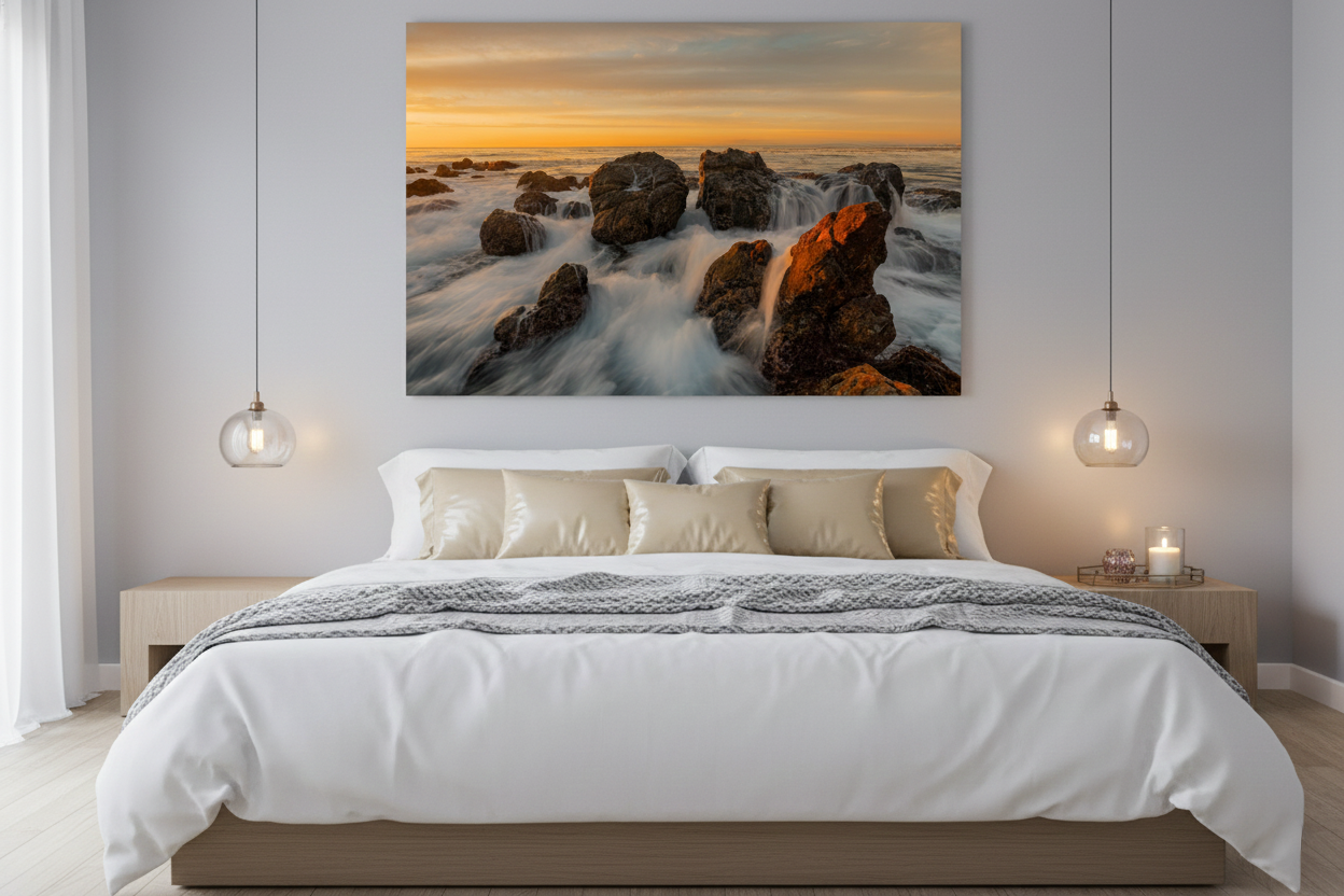 Frameless acrylic print of a California rocky coast sunset mounted above a king bed with beige pillows and pendant lights.
