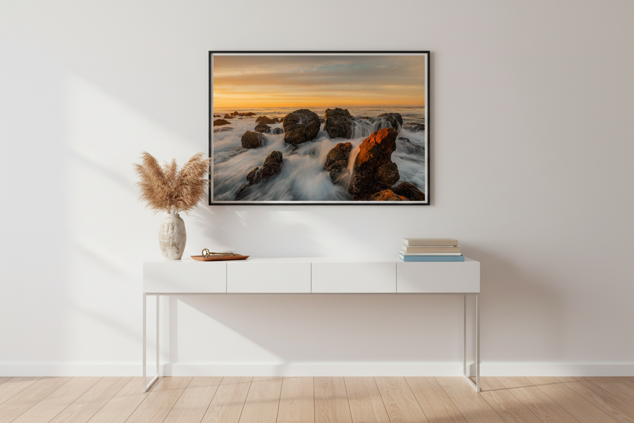 Framed California rocky coast seascape photograph mounted on a neutral wall above a sleek white console table.
