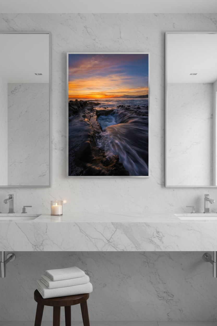 Large coastal ocean sunset print hanging between two mirrors on a white marble bathroom vanity wall.