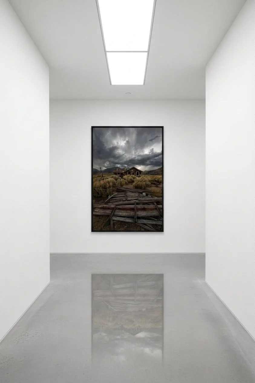 Fine art print of an abandoned cabin under storm clouds in the Eastern Sierra, framed on a white gallery wall at the end of a minimalist corridor with polished concrete floors.