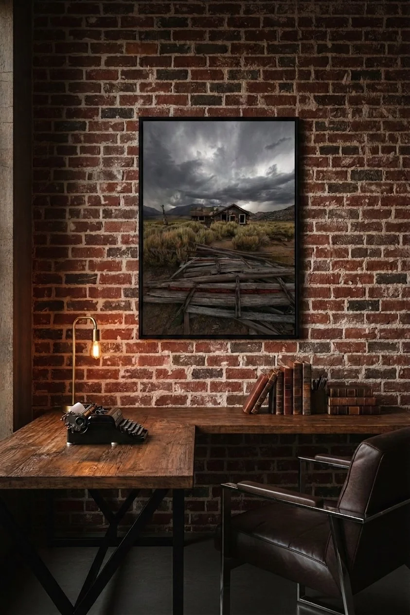 Fine art print of an abandoned Sierra cabin under dramatic storm clouds hanging on an exposed brick wall over a rustic wooden desk with a vintage typewriter.