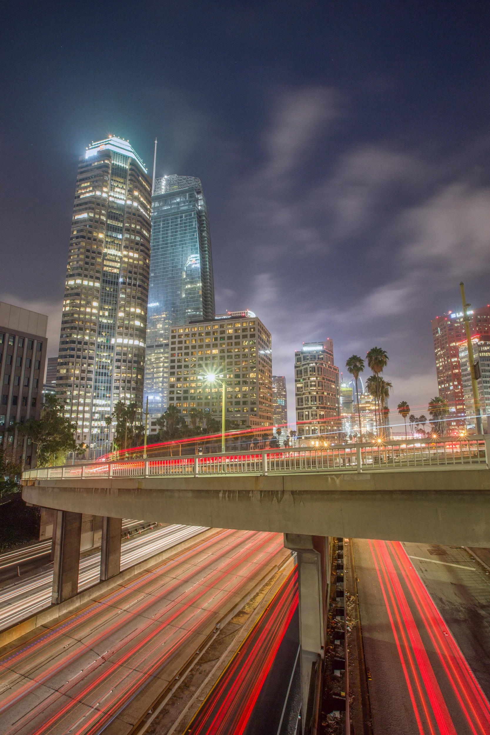 Long exposure of traffic light trails streaming along the 110 Harbor Freeway beneath Downtown Los Angeles skyscrapers by Chris Ewen Crosby.