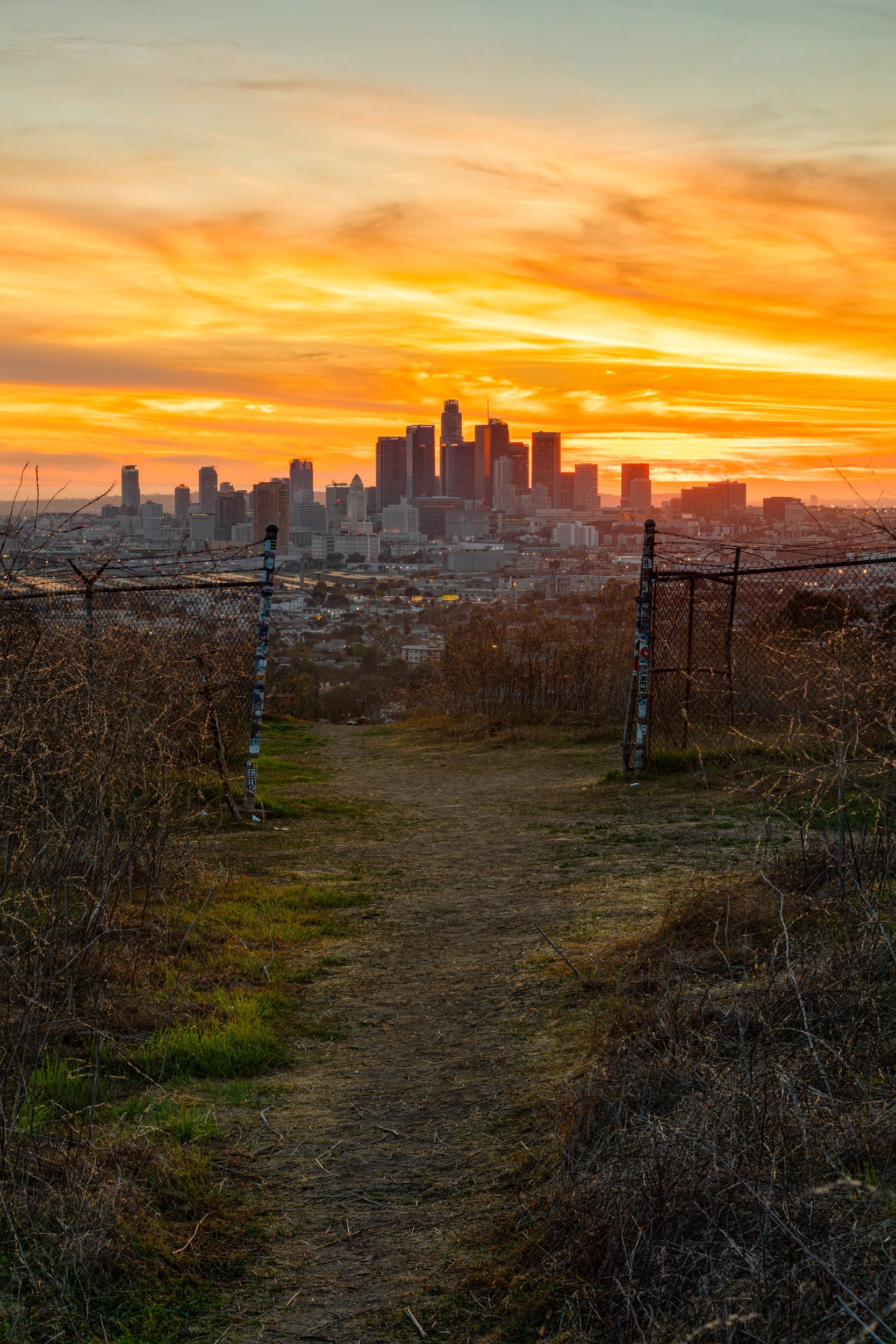 Dirt trail leading toward a glowing orange sunset behind the Downtown Los Angeles skyline at Ascot Hills Park by Chris Ewen Crosby.