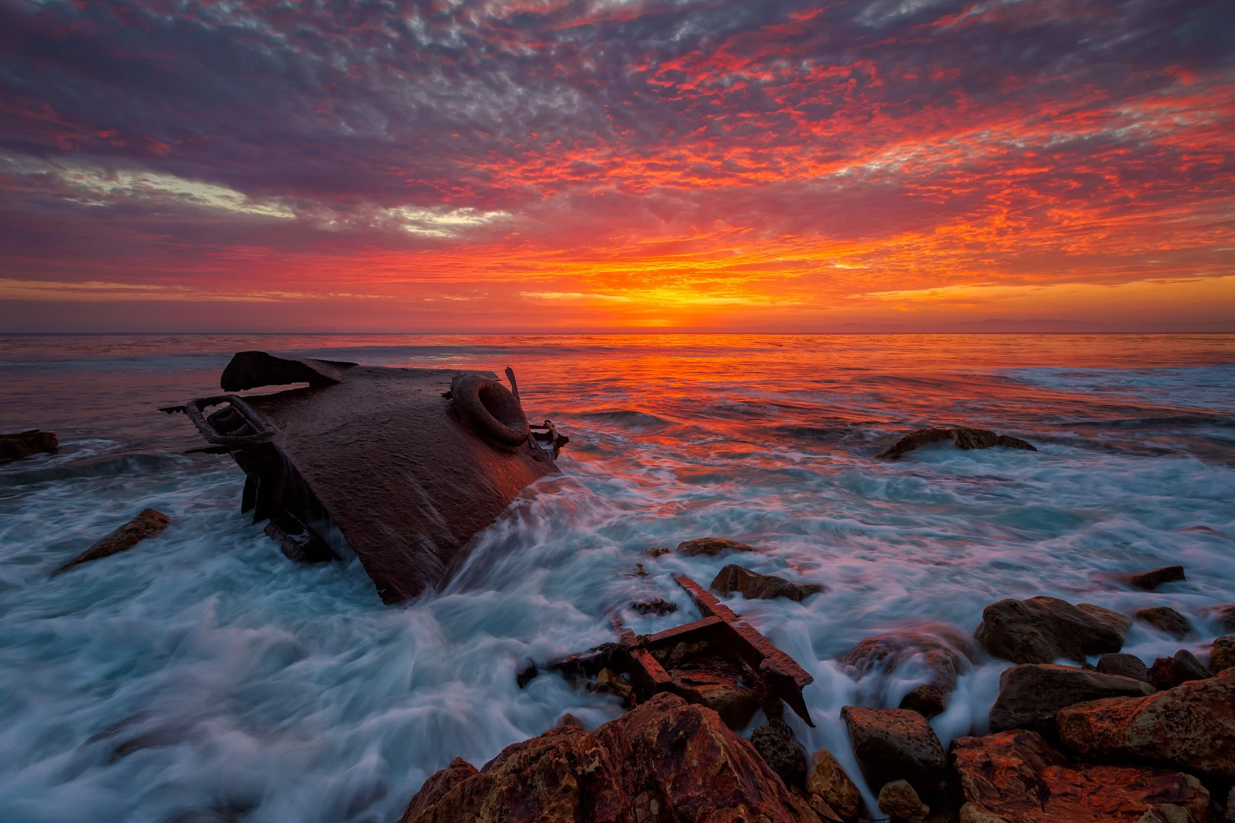 Horizontal shot of waves crashing over the rusting iron hull of the SS Dominator shipwreck in Palos Verdes under a blazing red sunset by Chris Ewen Crosby.